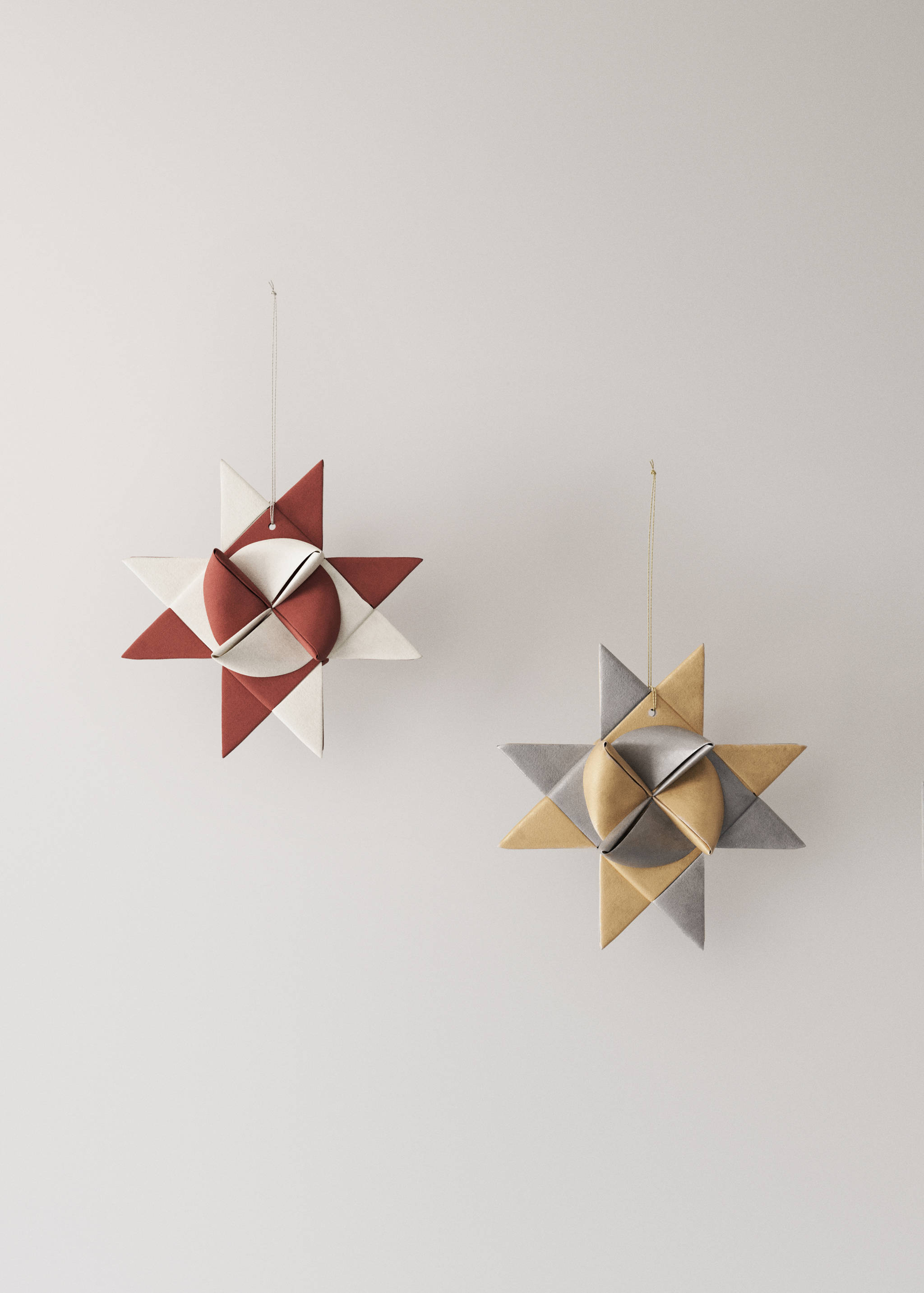 Two geometric star-shaped Christmas ornaments hanging on strings, one in terracotta and off-white, one in silver and gold.