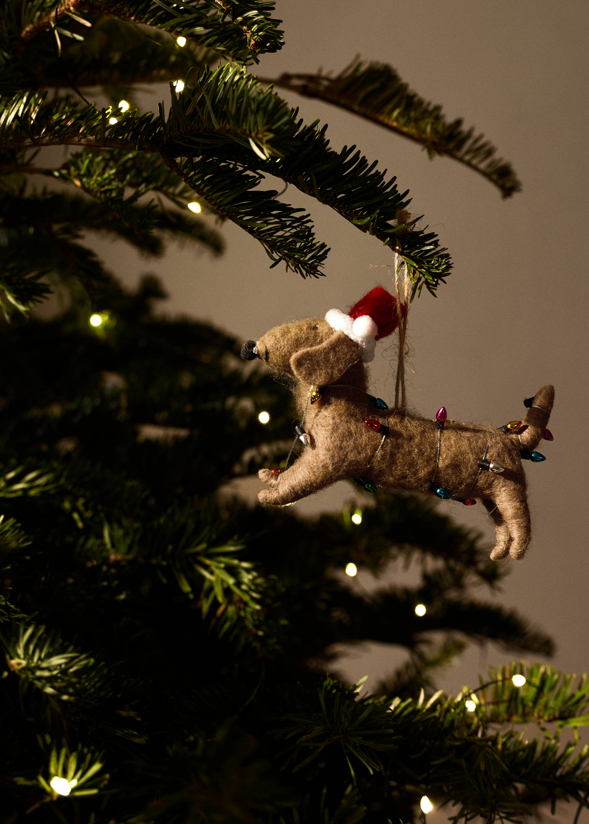 Brown felt dachshund Christmas ornament wearing a Santa hat and wrapped in fairy lights.
