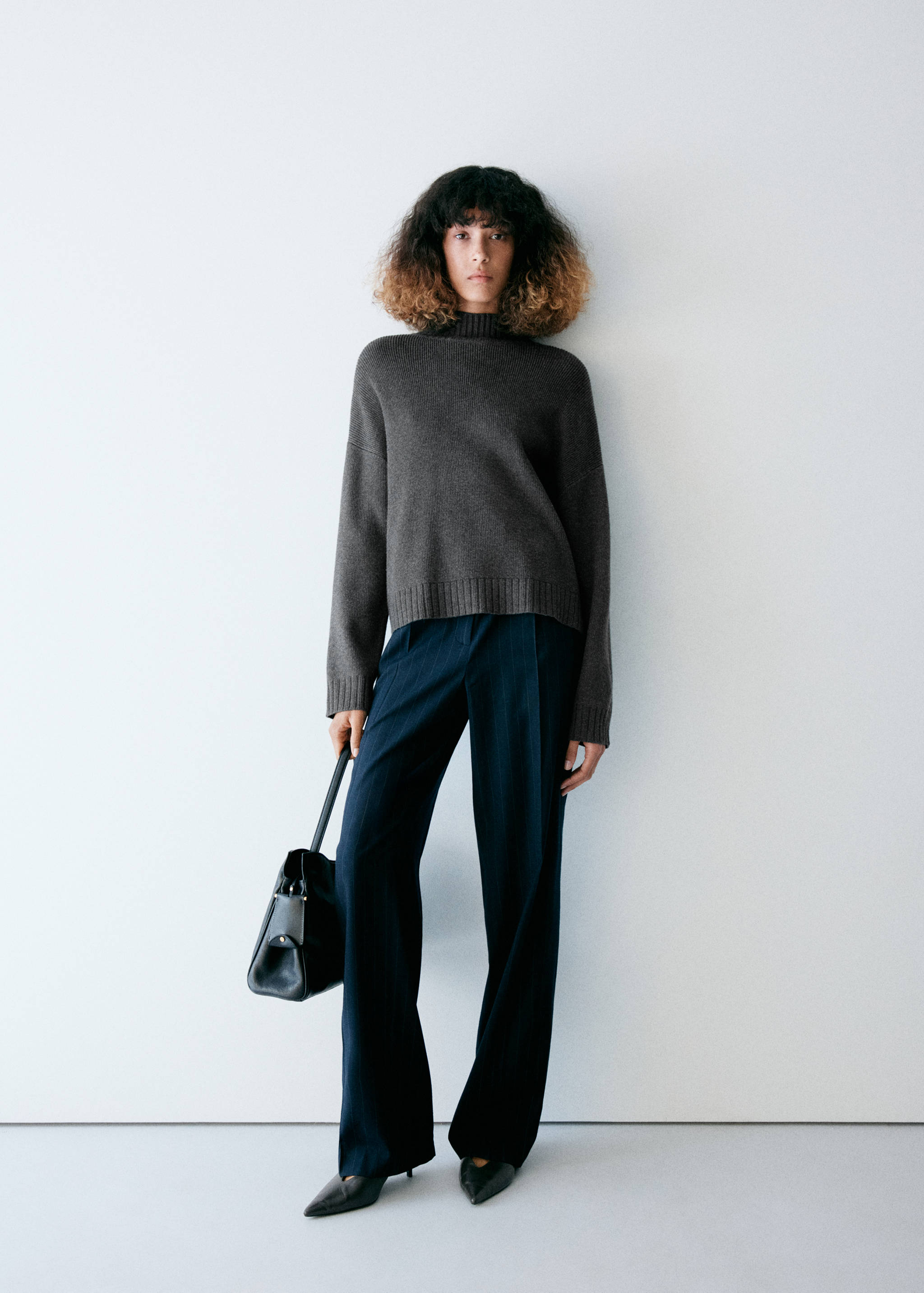 Dark grey knitted jumper with a high ribbed collar and dropped shoulders, worn with trousers and a handbag.
