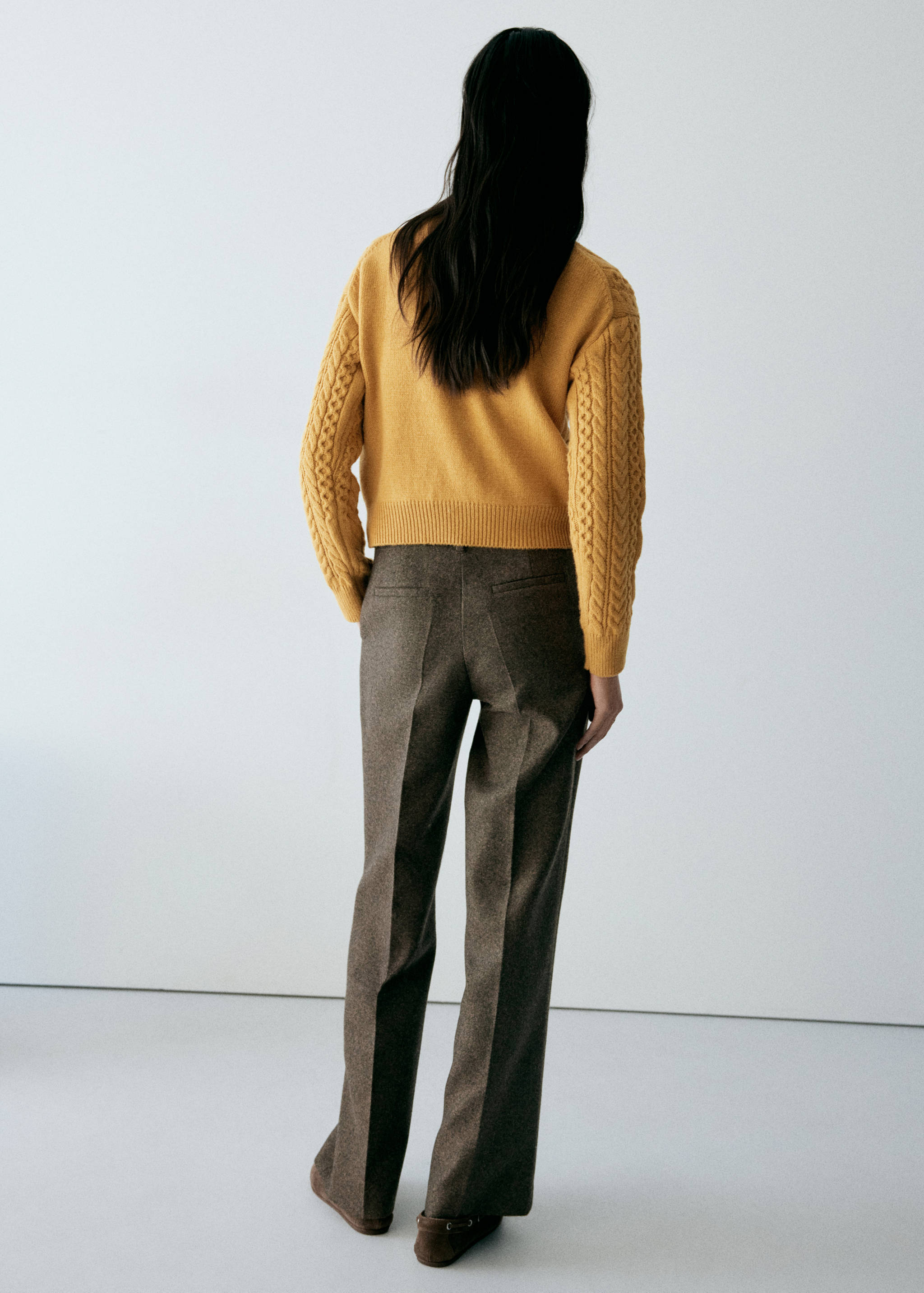 Mustard yellow cable-knit jumper from the back, worn with brown trousers.