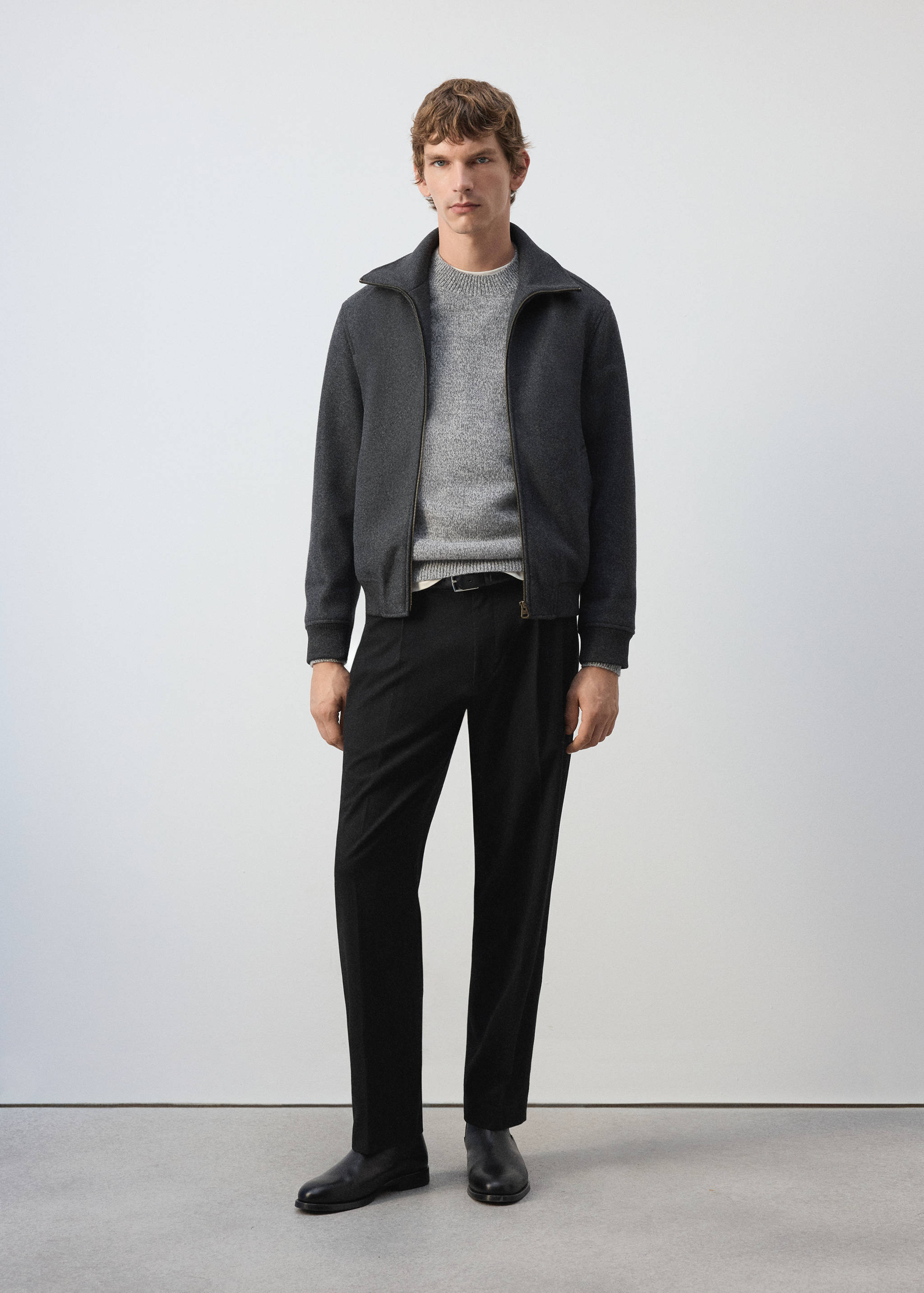 Dark grey zip-up jacket with a high collar and ribbed hem, worn with a grey sweater and black trousers.