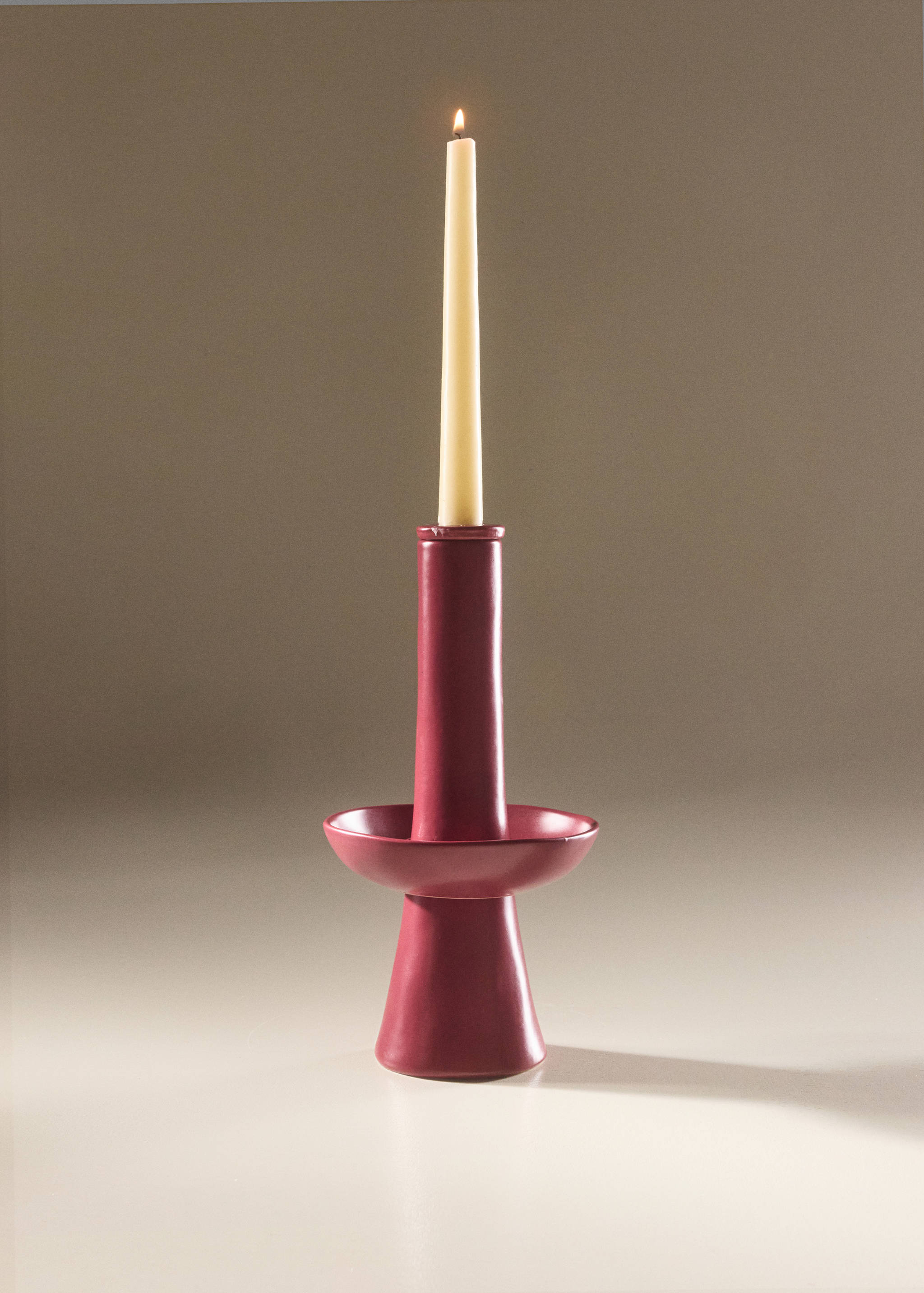 Burgundy ceramic candleholder holding a lit ivory taper candle.
