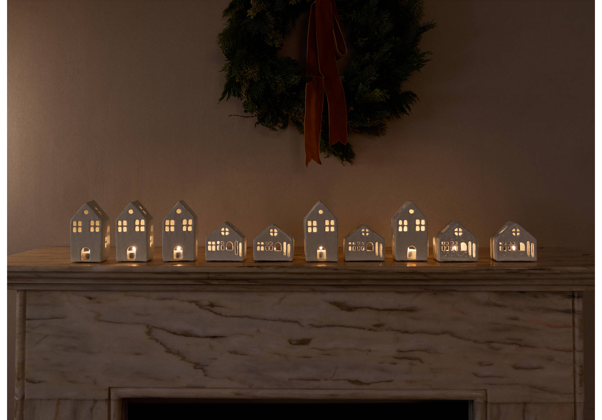 Ceramic house candle holders lined up on a mantelpiece, glowing with lit candles.