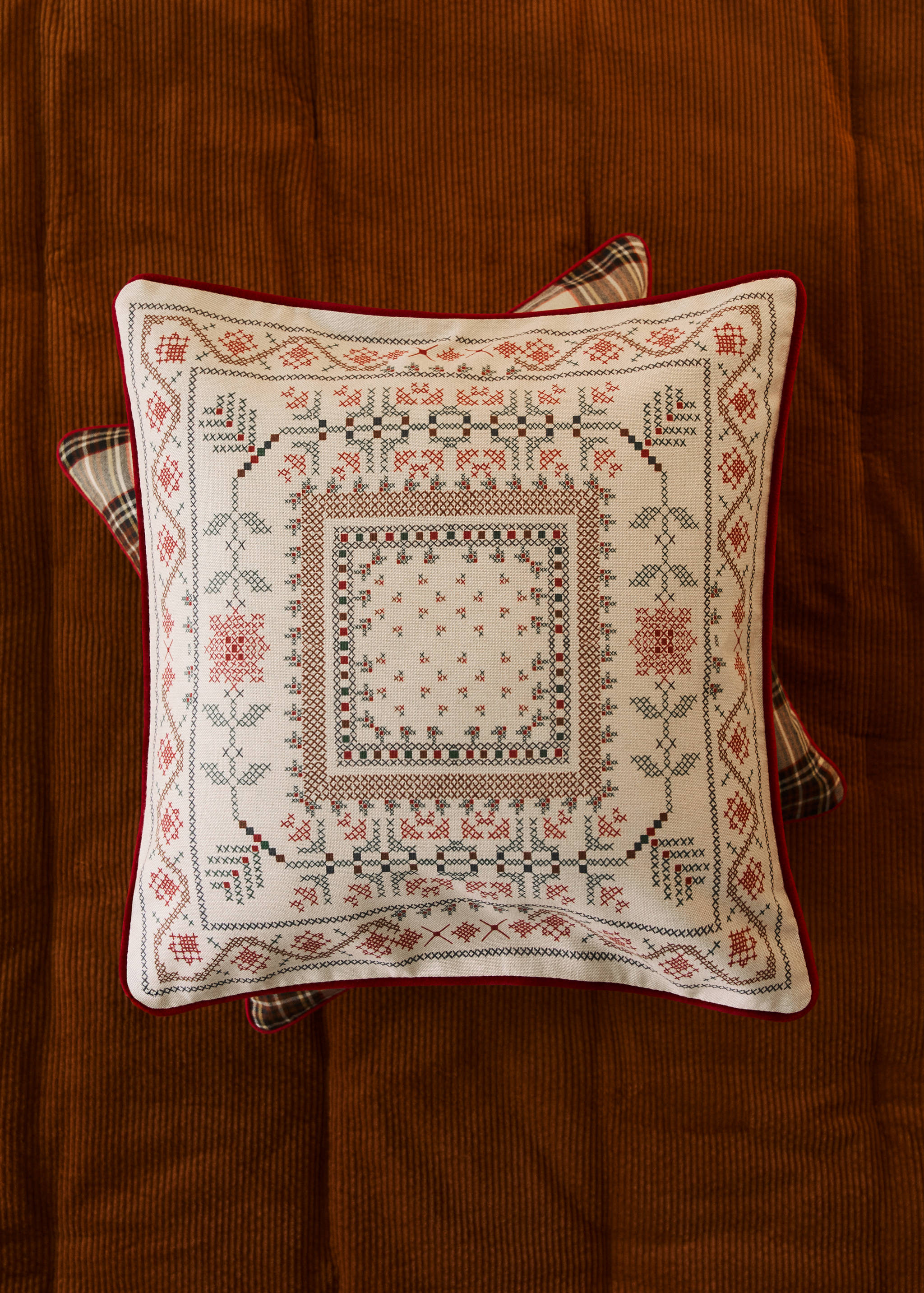 Off-white square cushion cover with red, green and brown cross-stitch embroidery and red piping.