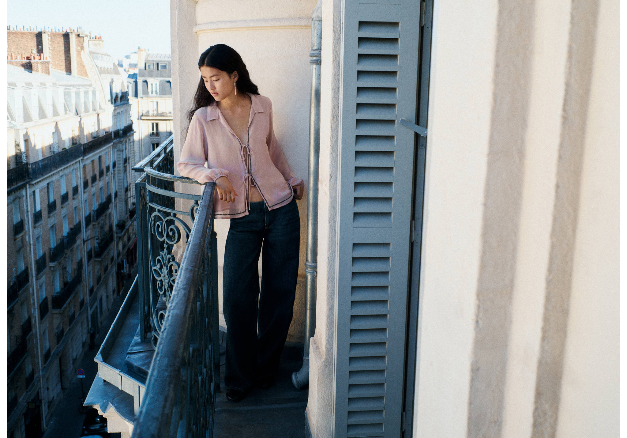 Pink semi-sheer blouse with front ties, worn with dark denim trousers on a balcony.
