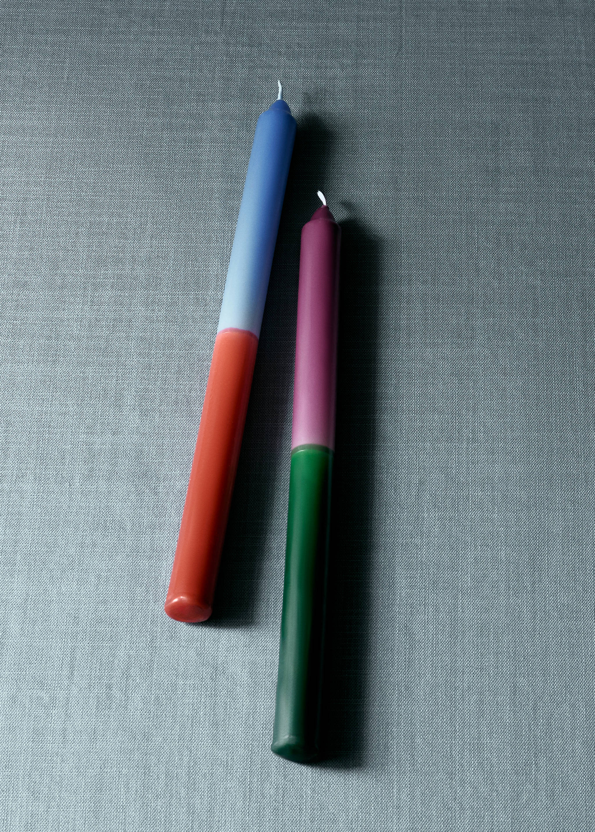 Two colour-blocked tapered candles lying on a textured grey fabric.