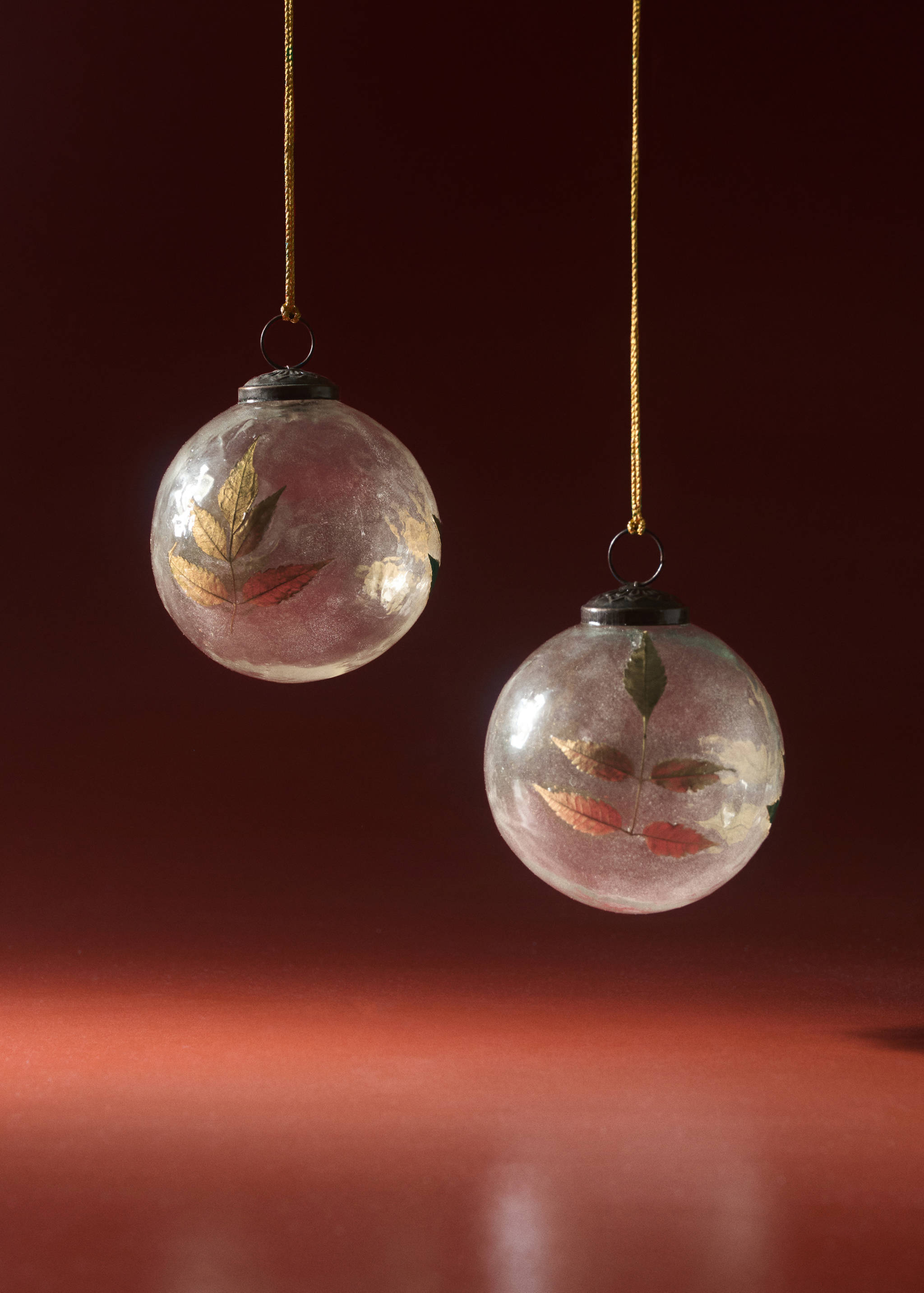 Two round glass Christmas ornaments containing pressed leaves, suspended by gold cords.