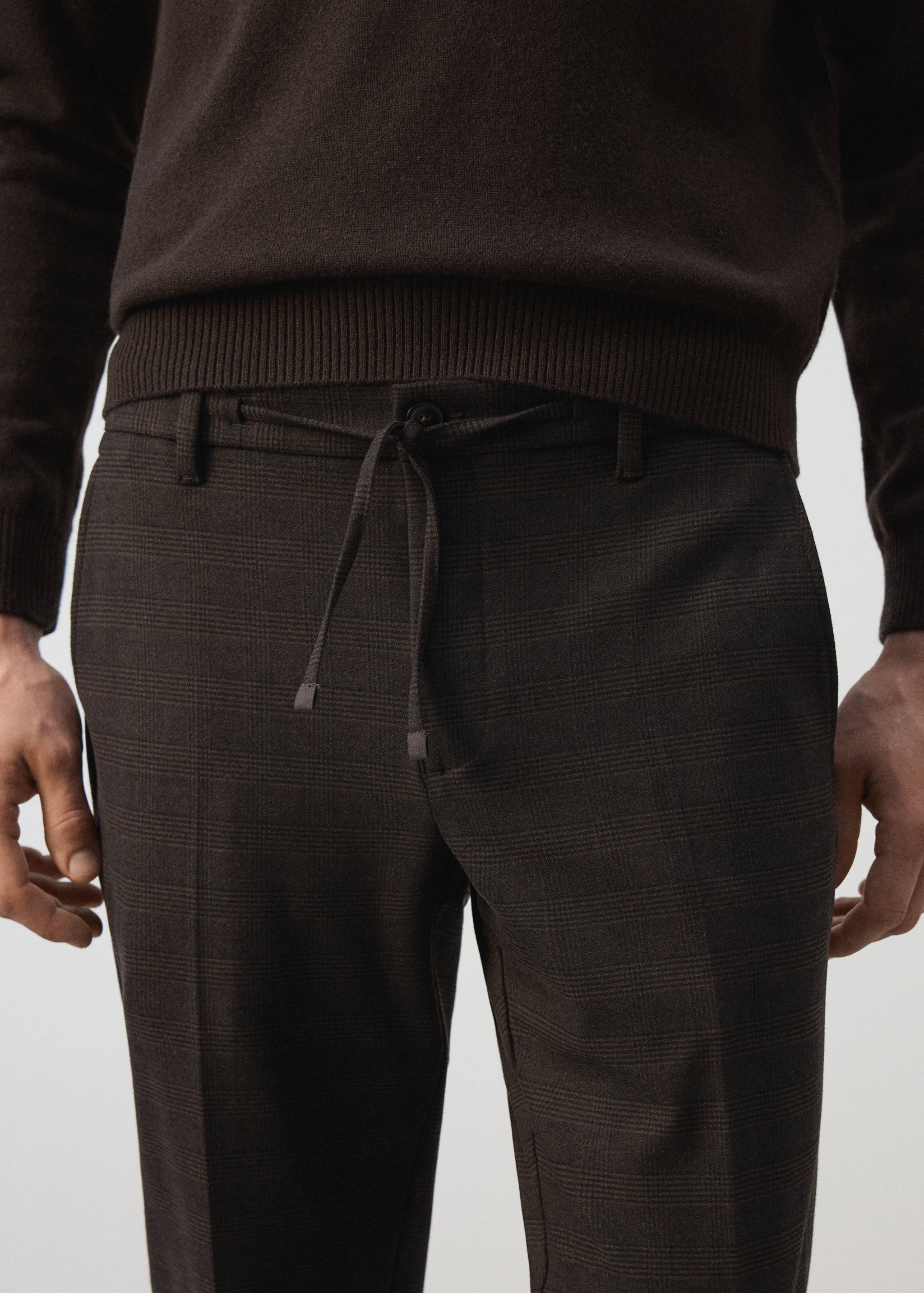 Dark checked trousers with a drawstring waist detail, paired with a dark brown jumper.