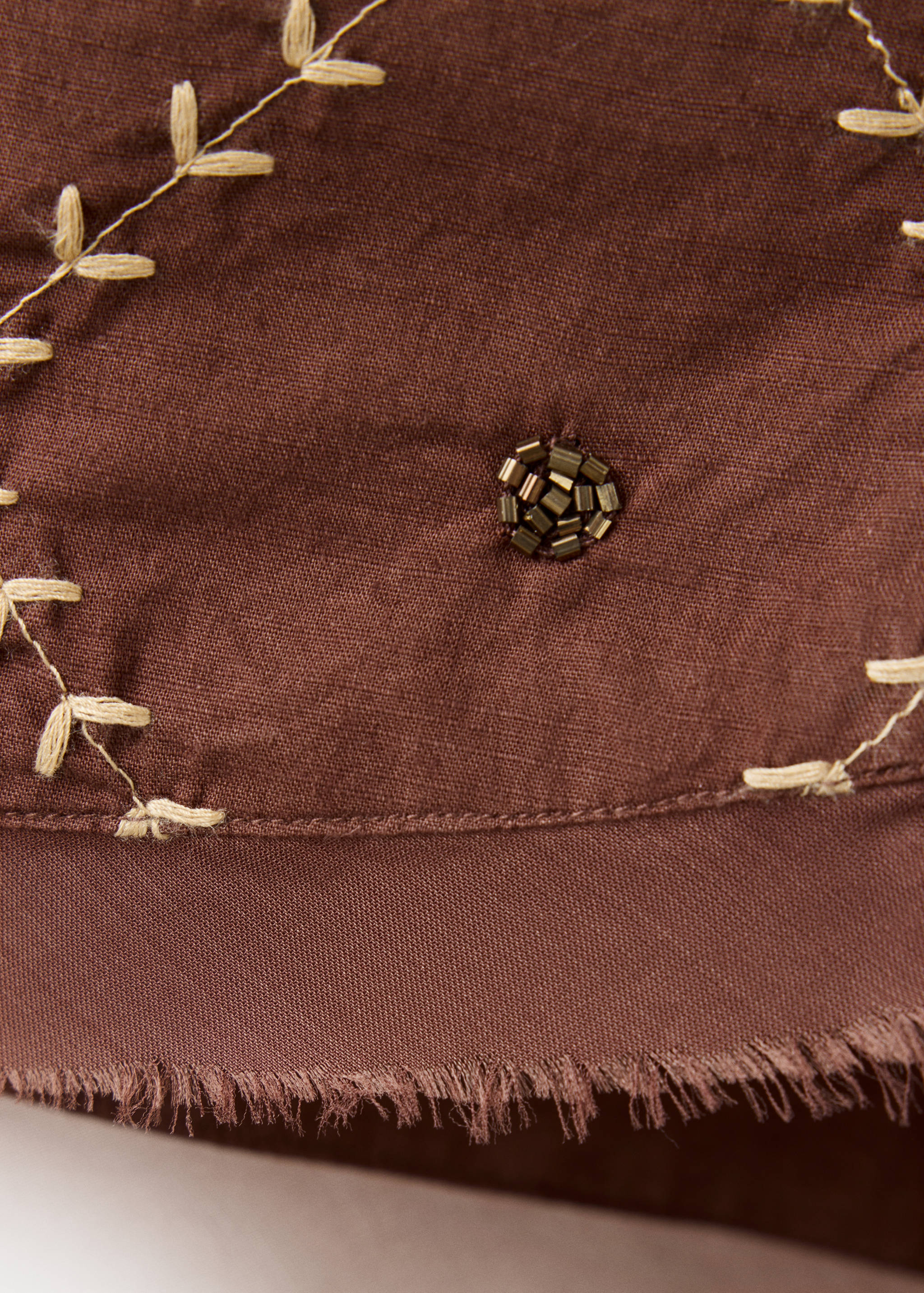 Close-up of a brown shirt fabric with decorative beige embroidery and bead details.