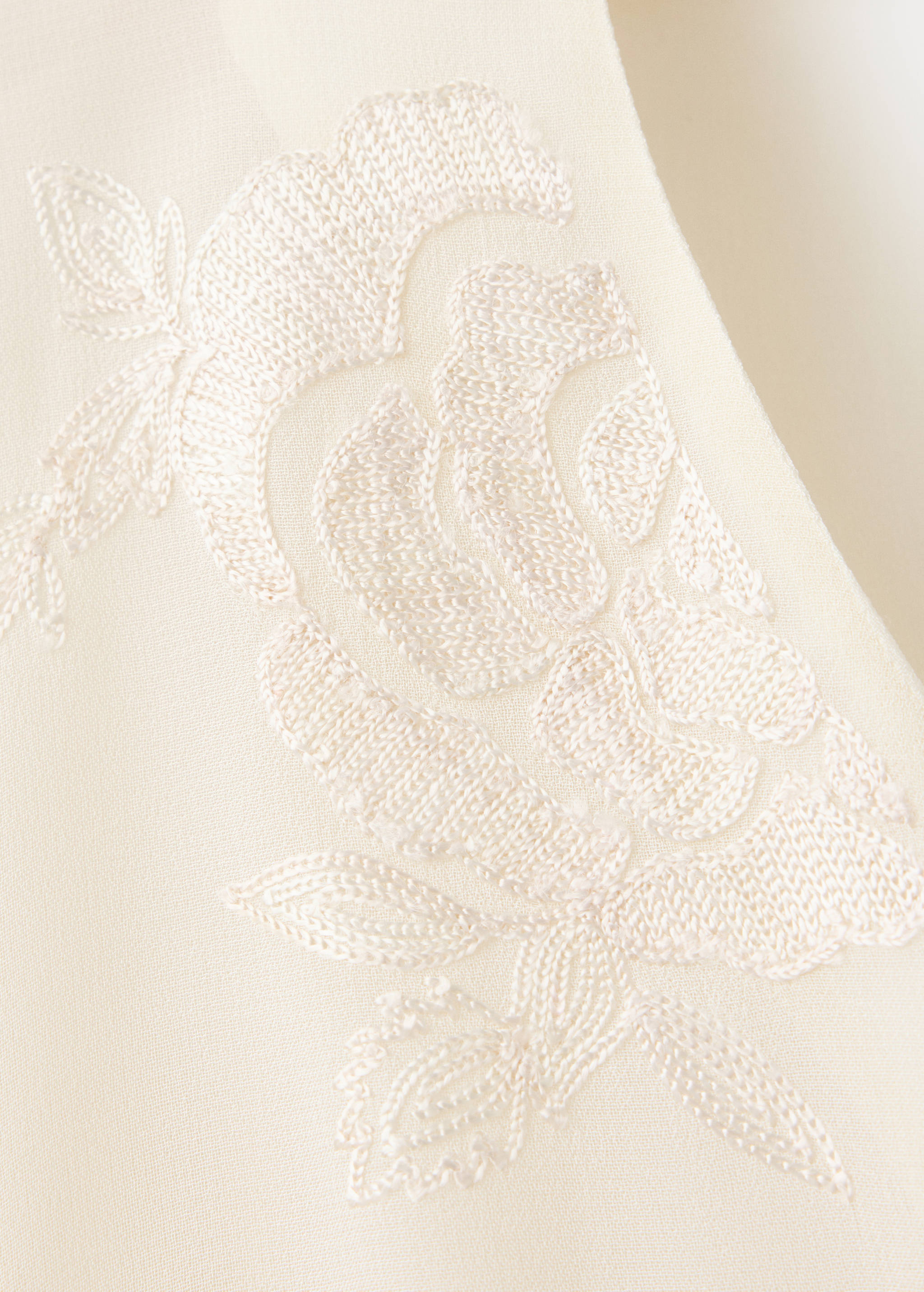 Primer plano del tejido de la blusa en color crudo con bordados florales tonales.