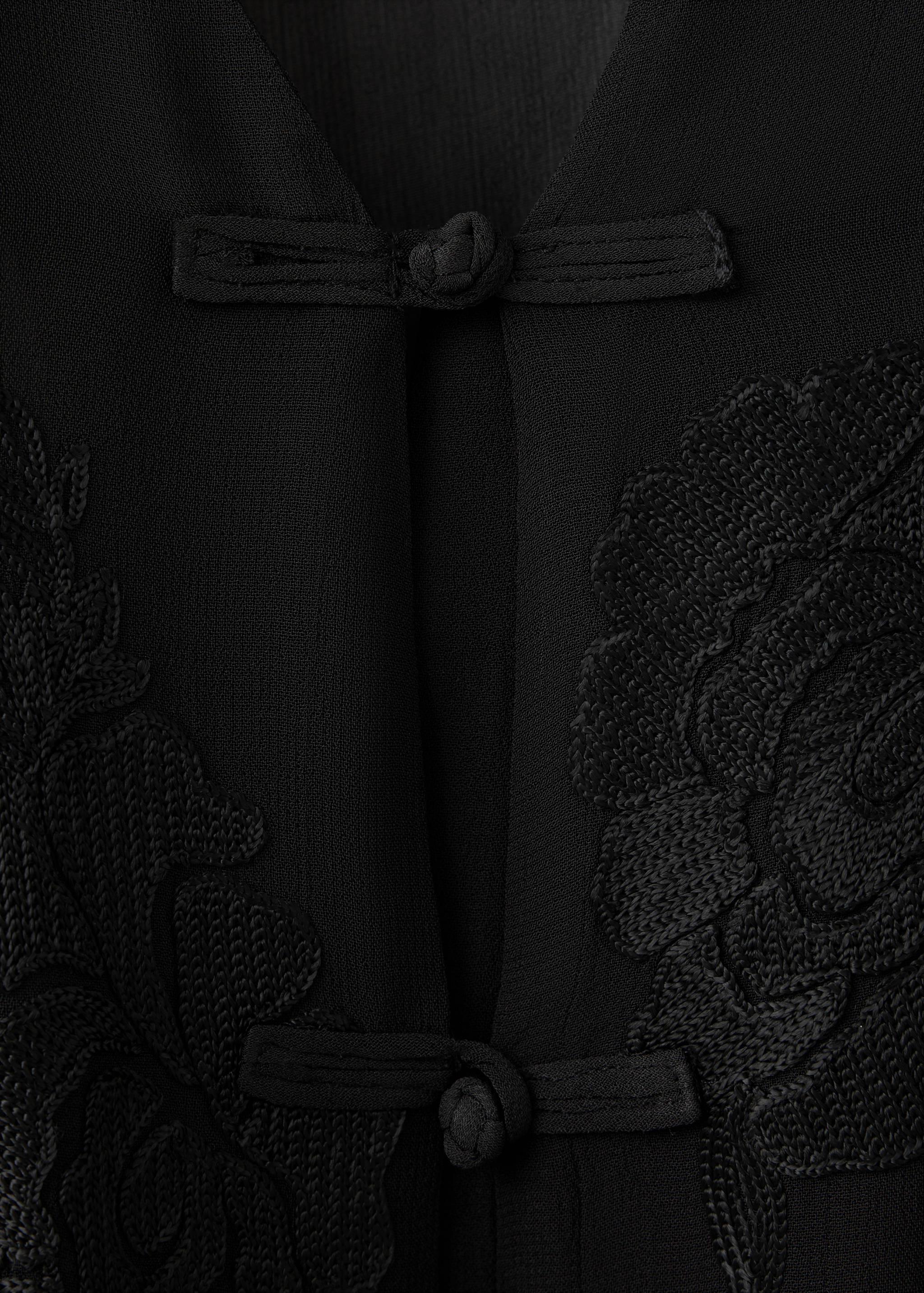 Primer plano de una blusa negra con pasamanería al tono y detalles florales bordados.