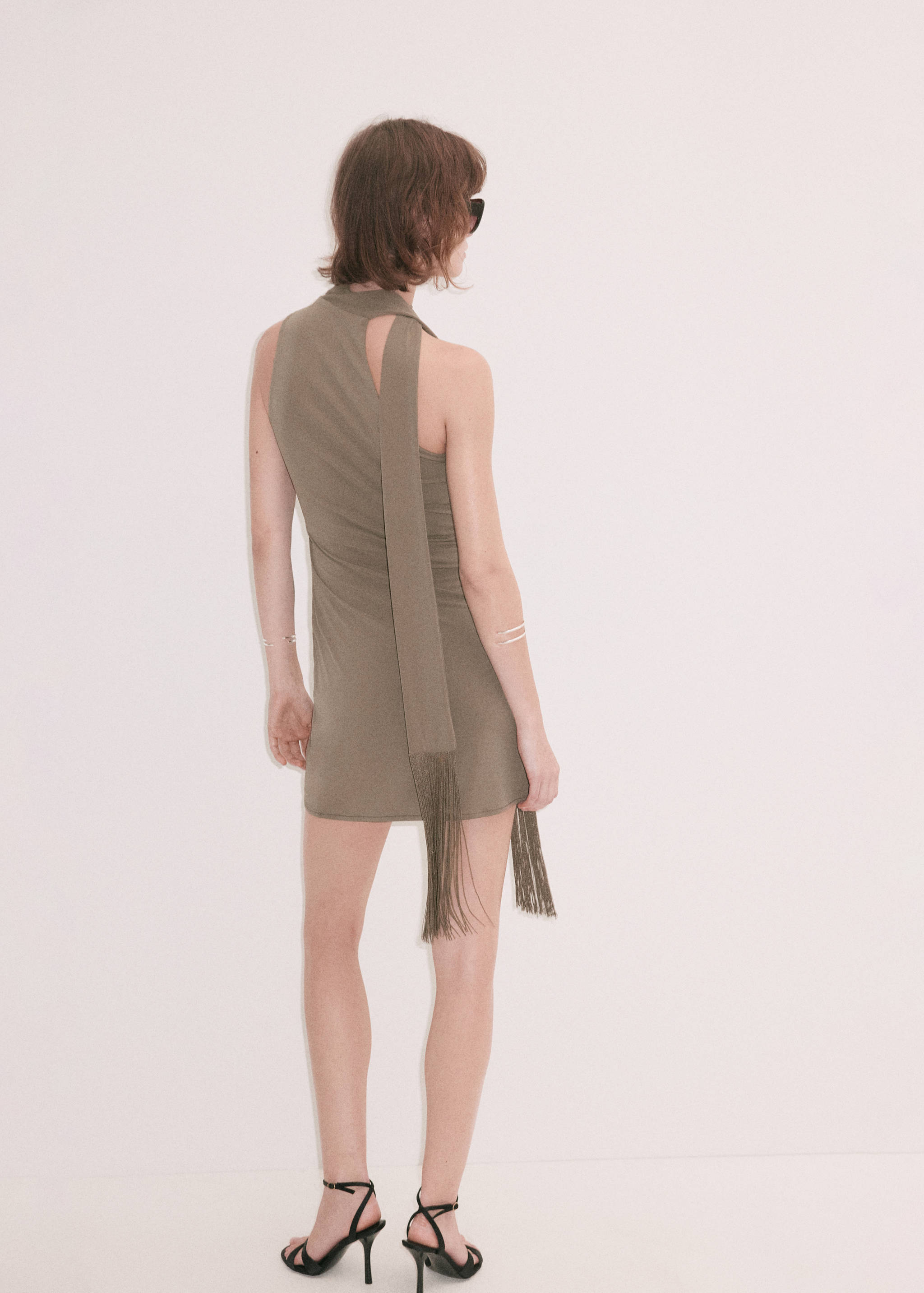 Khaki mini dress seen from the back, featuring a sleeveless design and a fringed scarf detail.