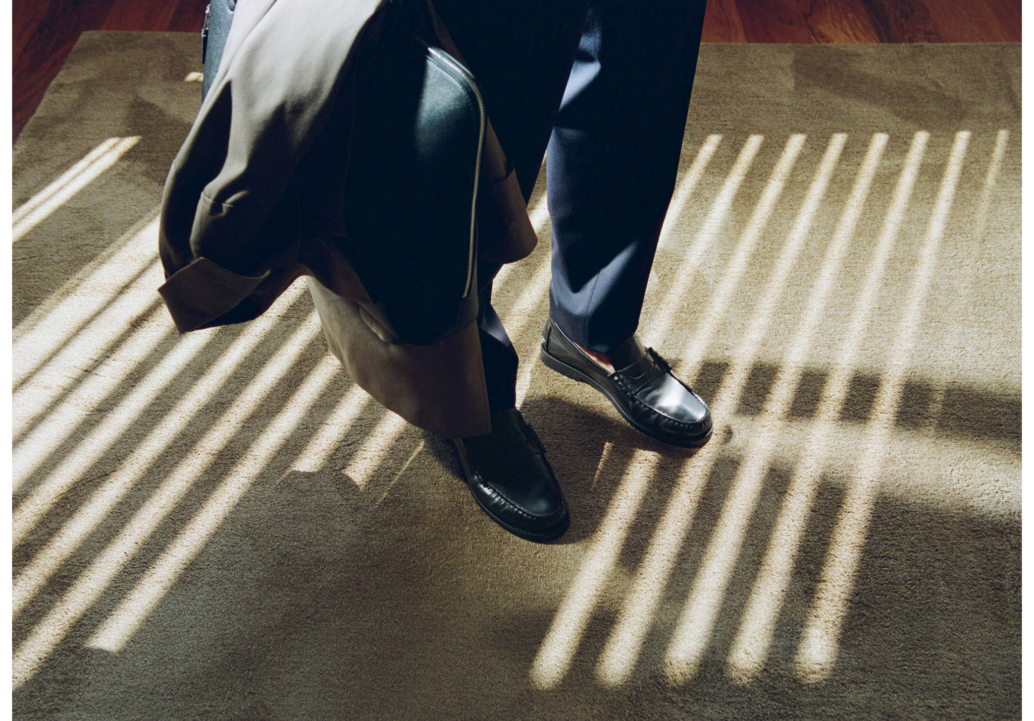 Mocasines penny de piel negra con pantalones oscuros sobre una alfombra.
