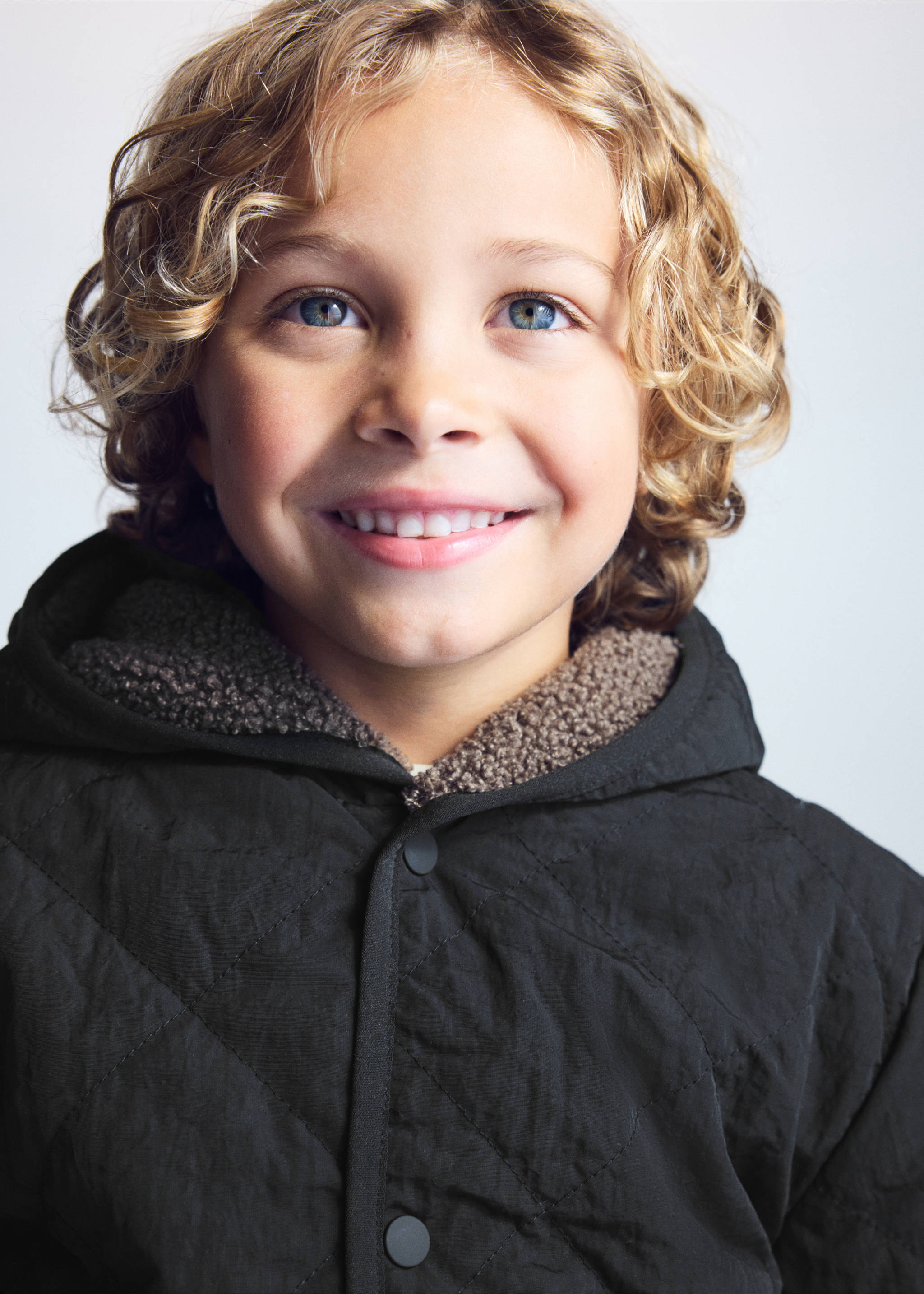 Close-up of a child wearing a black quilted anorak with a hood and textured faux-fur lining., Black. Ref: 37025153-00.
