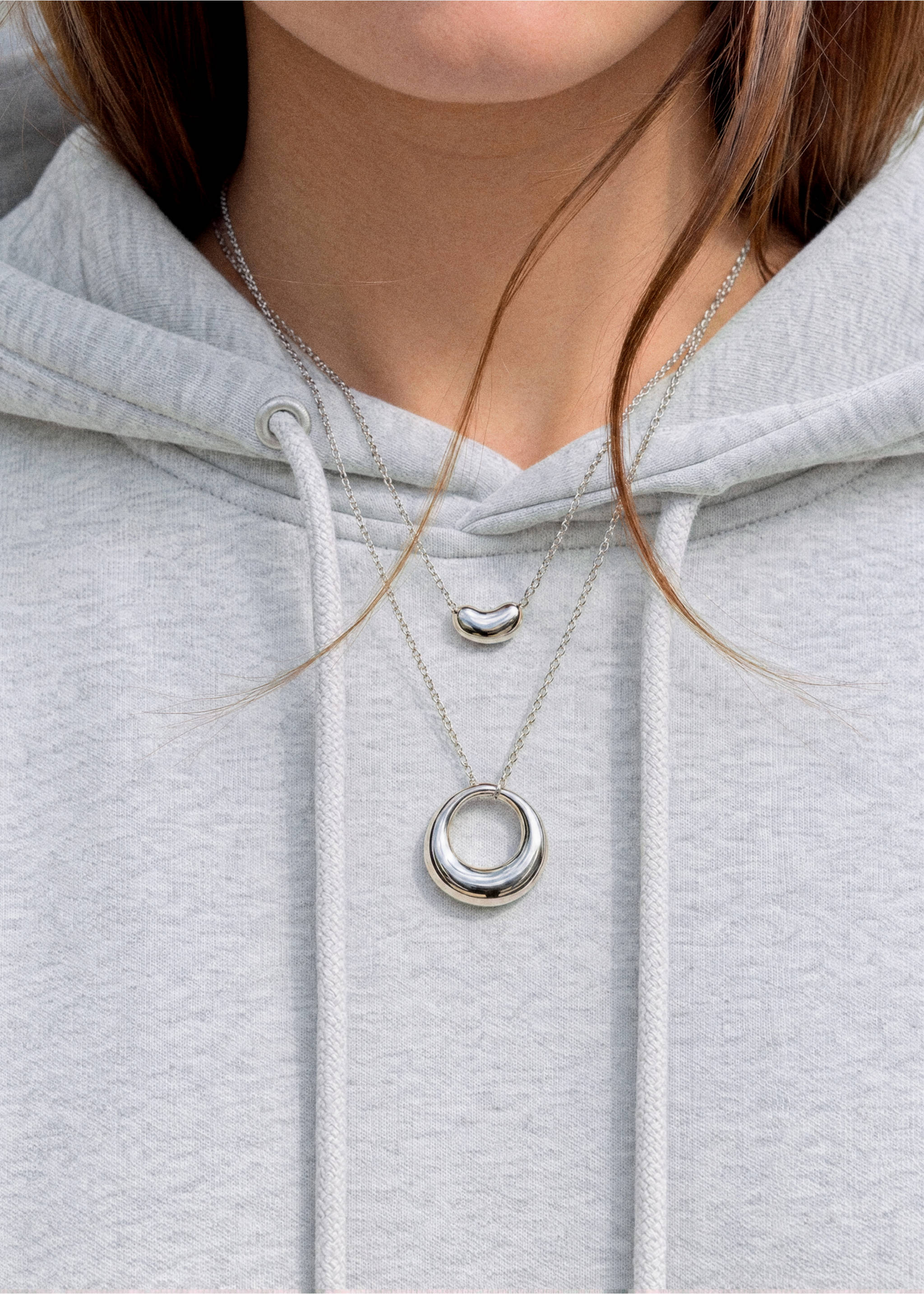 Dos collares plateados con colgantes en forma de judía y circular, combinados con una sudadera gris., Plata. Ref: 37025187-00.