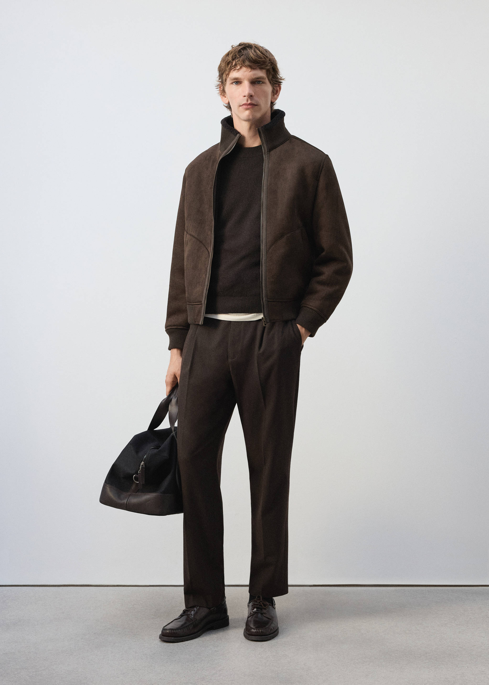 Dark brown zip-up jacket with a high neck, styled with a matching jumper, trousers, and a travel bag.
