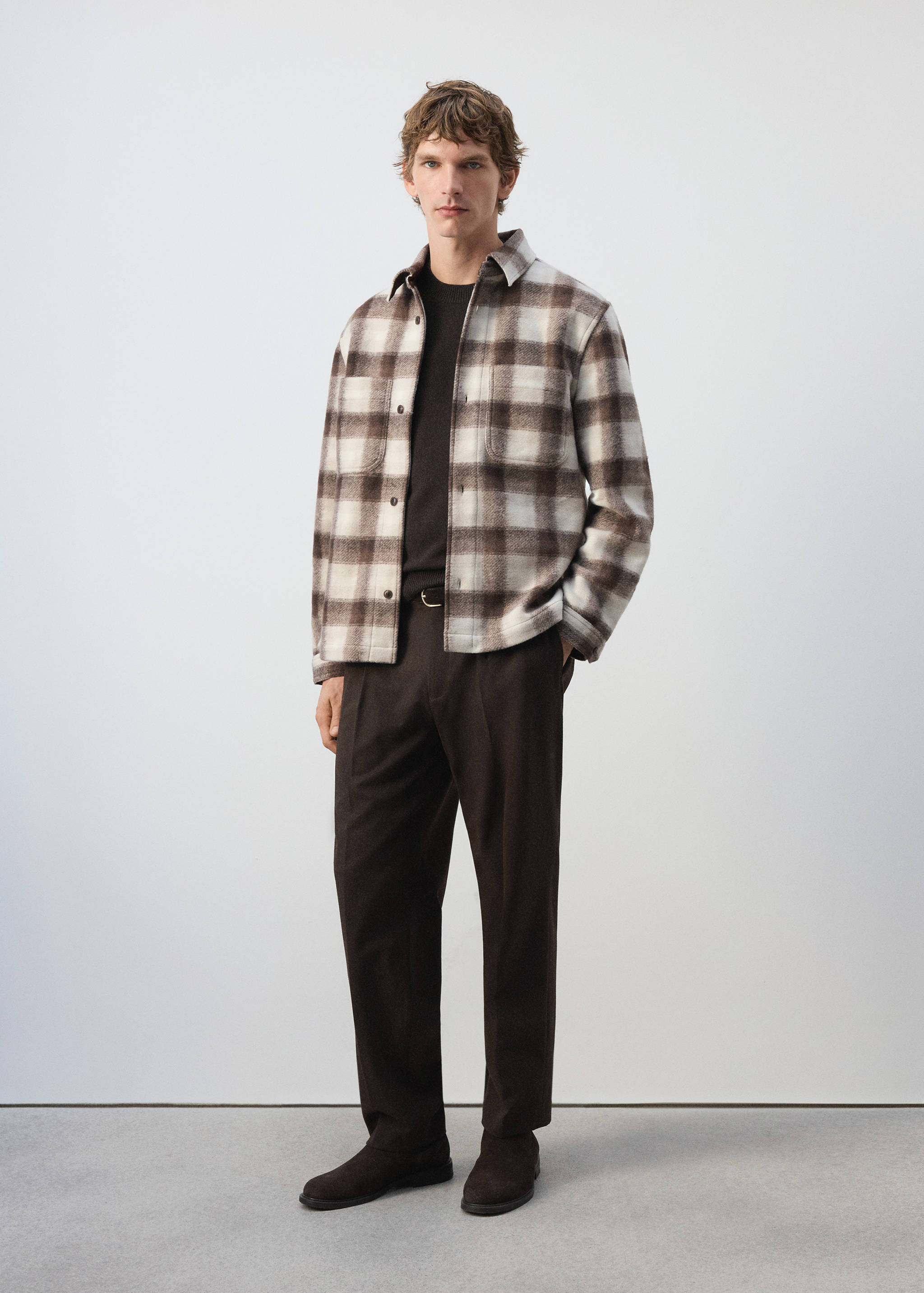 Sobrecamisa de cuadros en marrón y blanco llevada abierta sobre un jersey oscuro y pantalón.