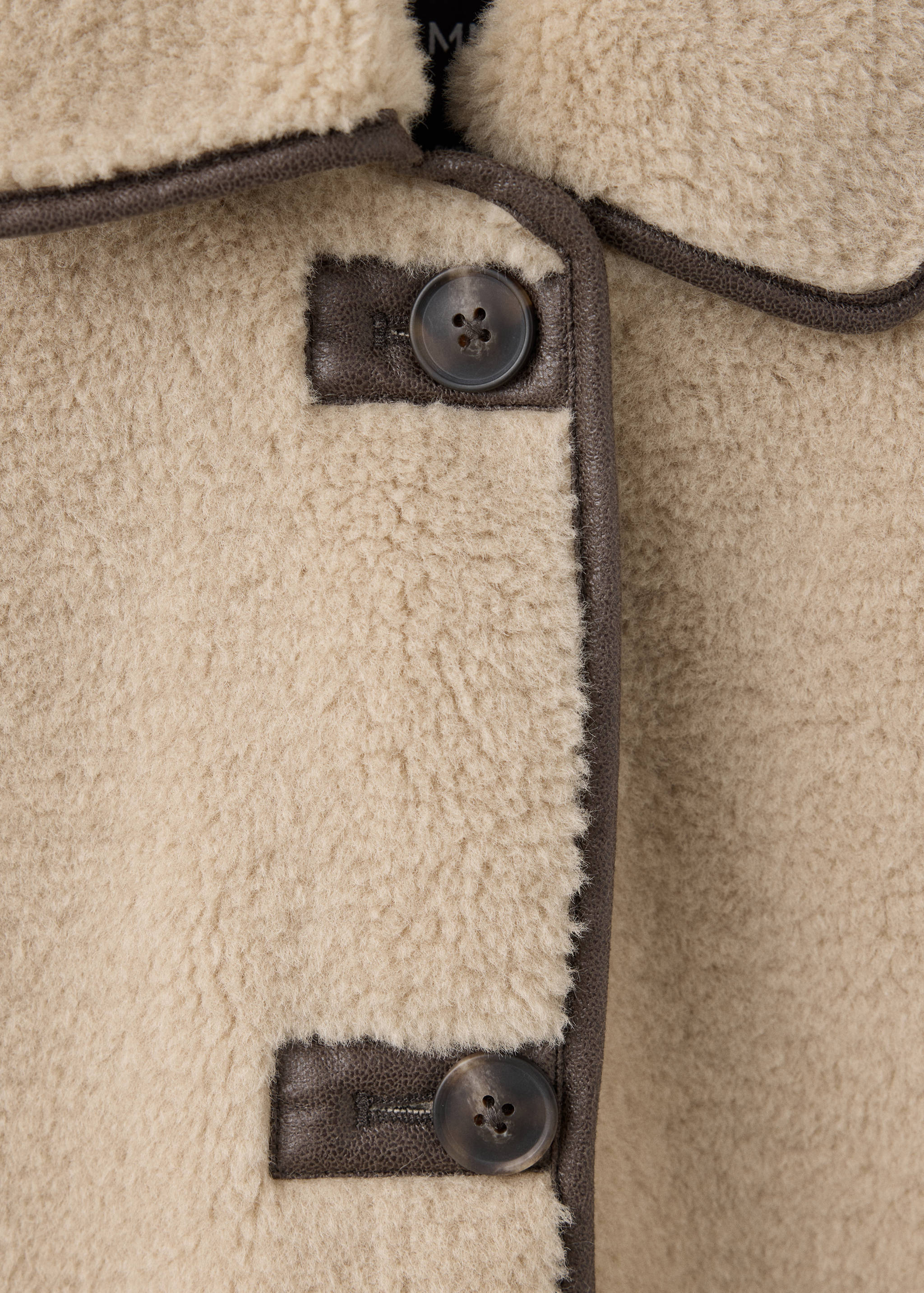 Detalle de abrigo beige de borreguillo sintético con ribetes y botones en marrón oscuro.