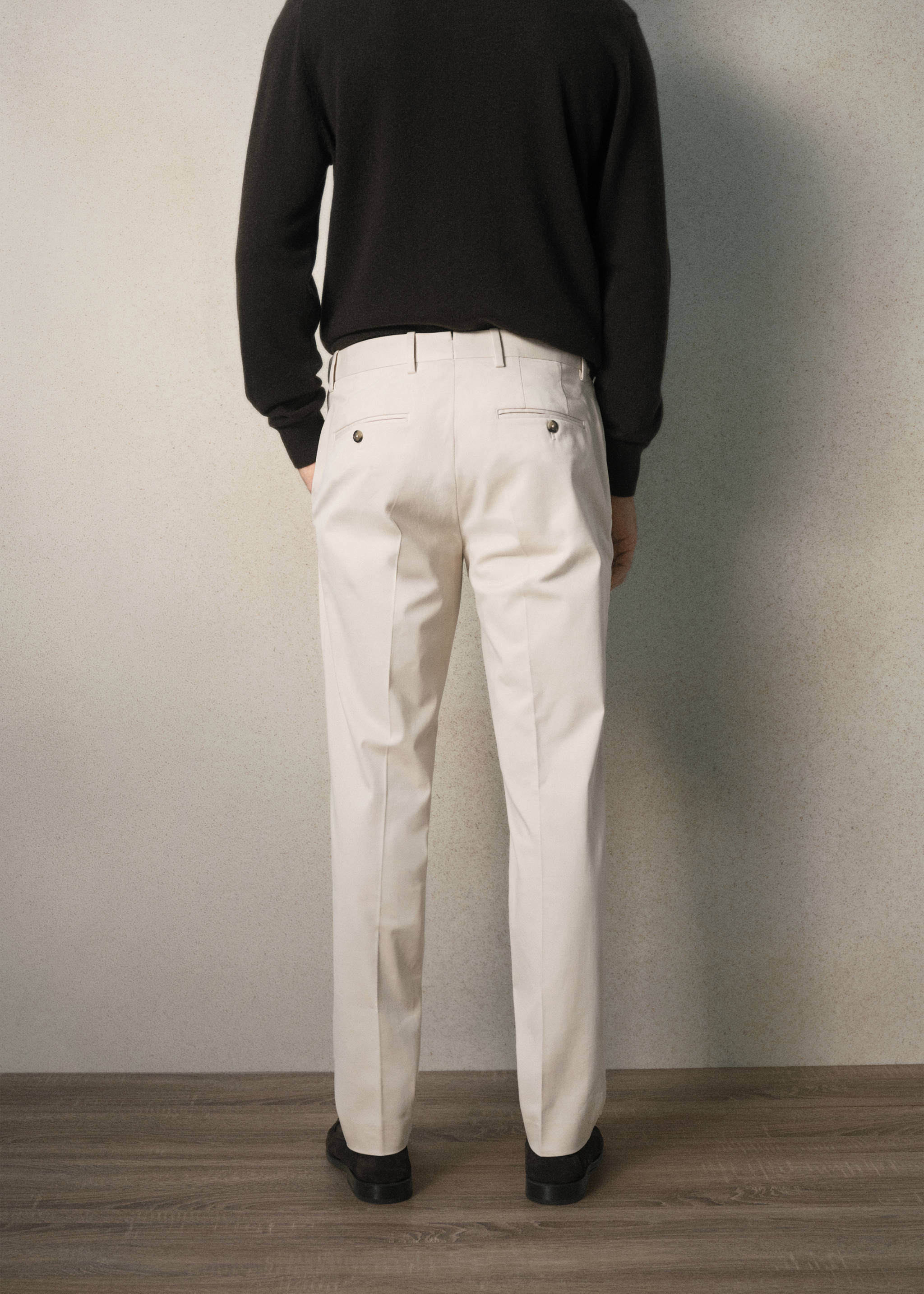Cream trousers seen from the back, featuring belt loops, pressed creases, and rear buttoned pockets.