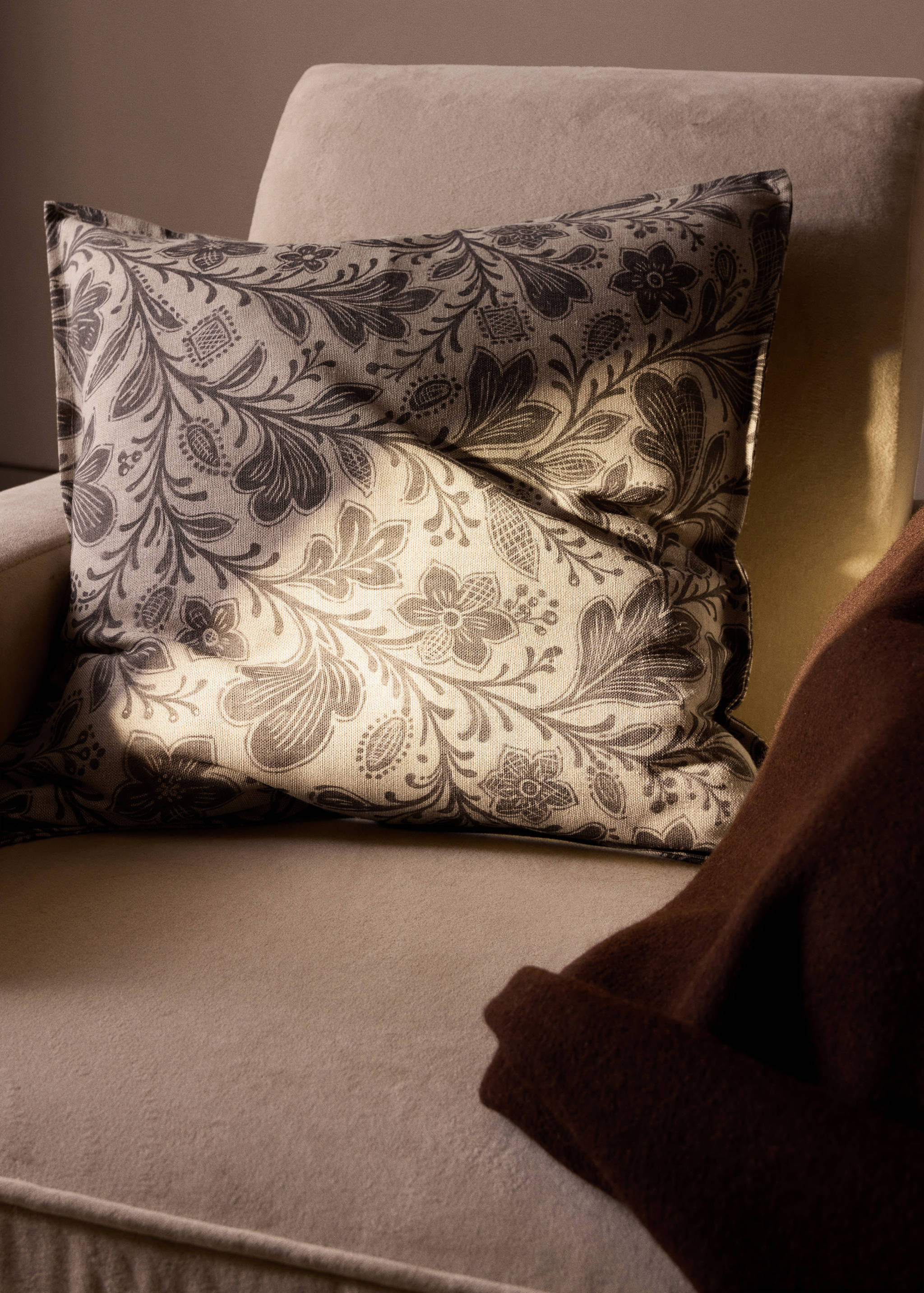 Square cushion cover with dark floral print on a light beige background resting on an armchair.
