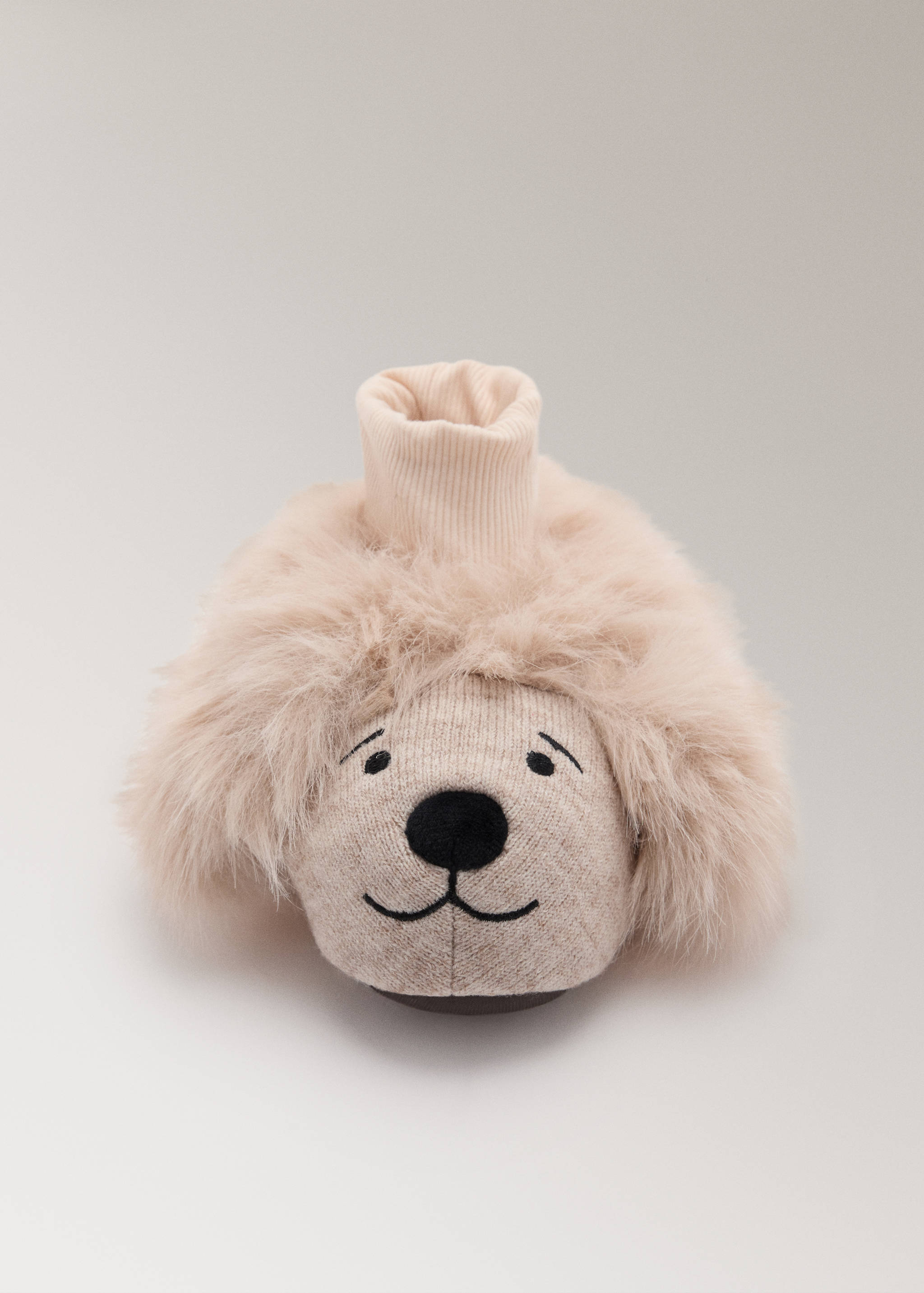 Primer plano de una zapatilla de casa infantil en color beige claro con forma de perro, ribete de efecto pelo mullido y detalles de la cara bordados.