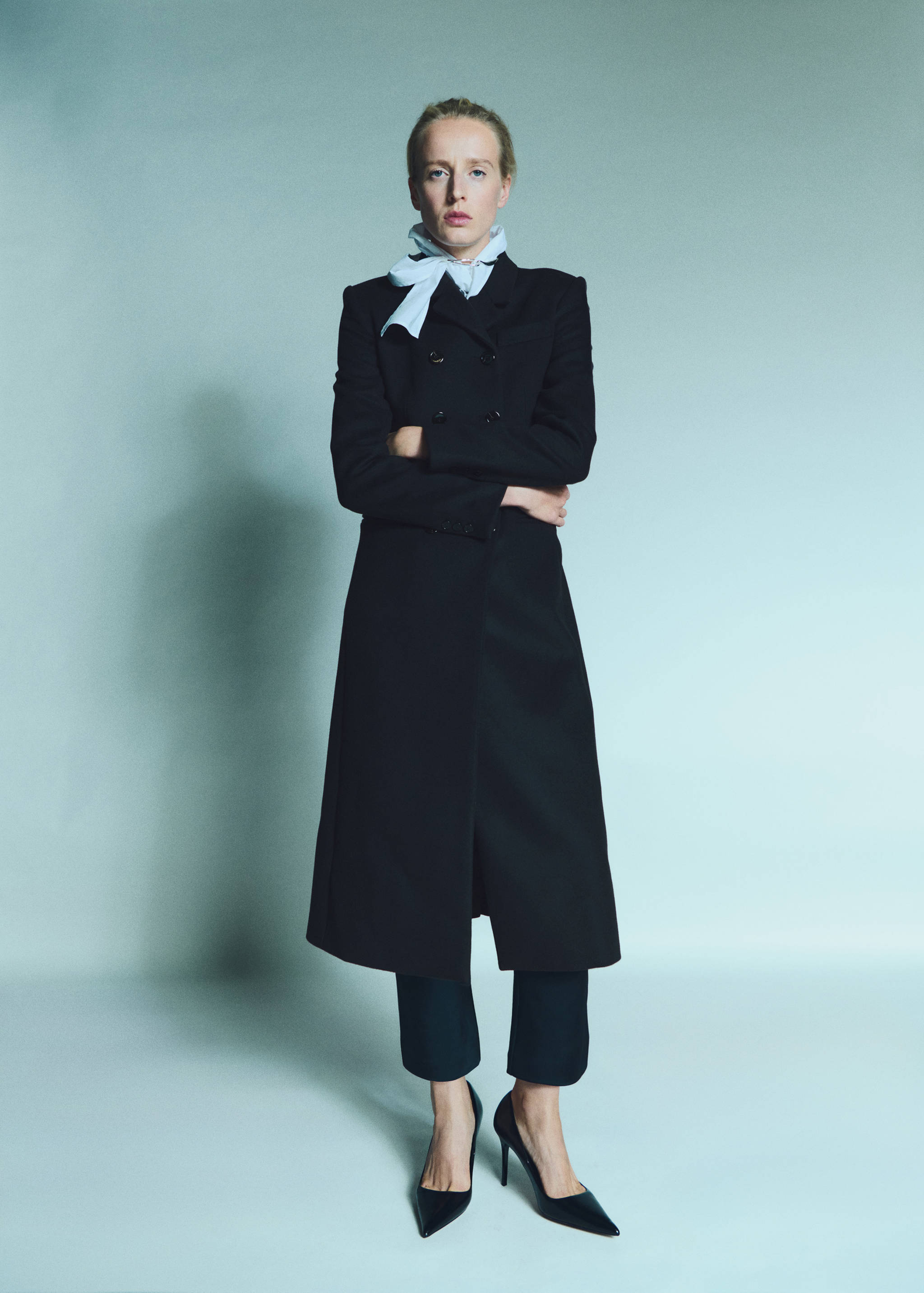 Long black double-breasted coat worn with cropped trousers and a light blue scarf.
