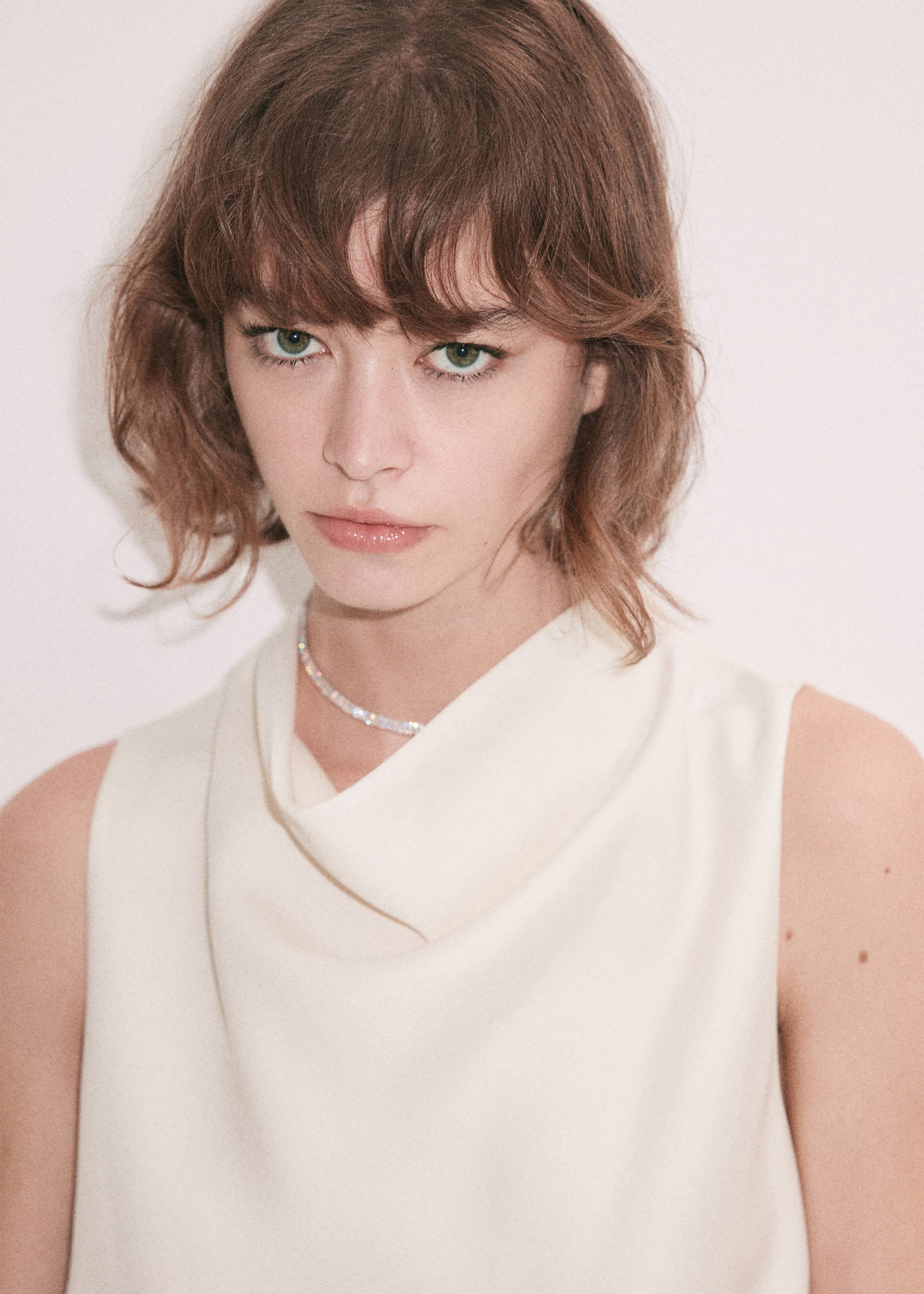 Close-up of a model's face wearing an off-white sleeveless top with a cowl neckline.