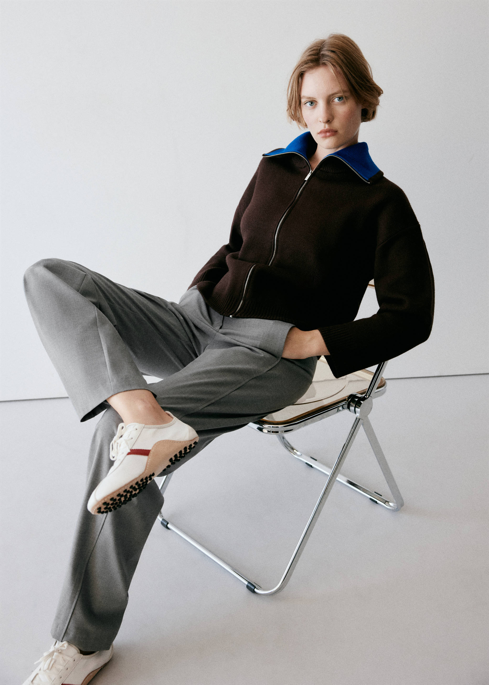Grey trousers styled with a brown zip-up jacket and retro sneakers.