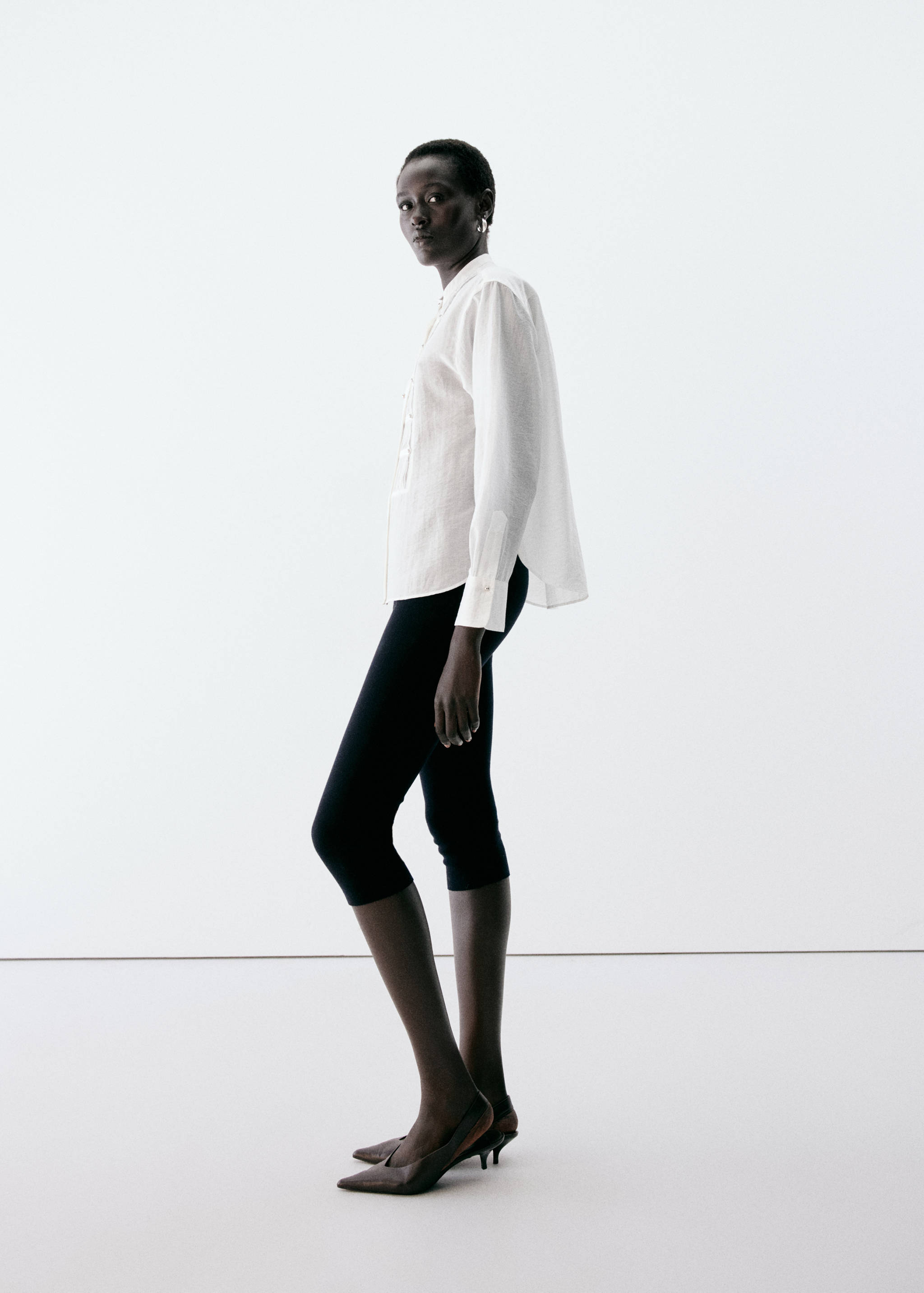 White long-sleeved button-up blouse with a relaxed fit and curved hem, worn with black cropped leggings.