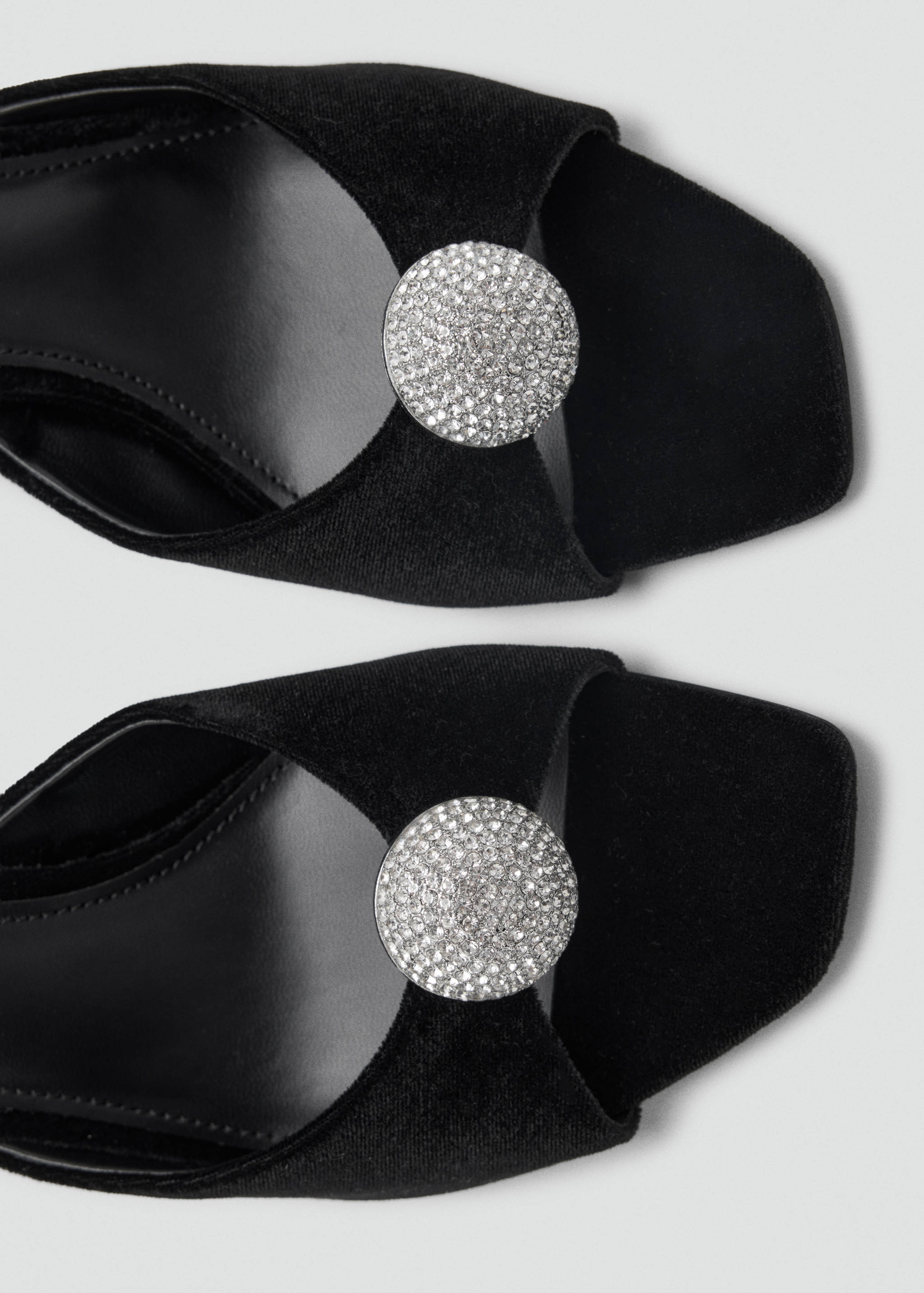 Close-up of black velvet sandals featuring a square toe and a circular rhinestone medallion detail.