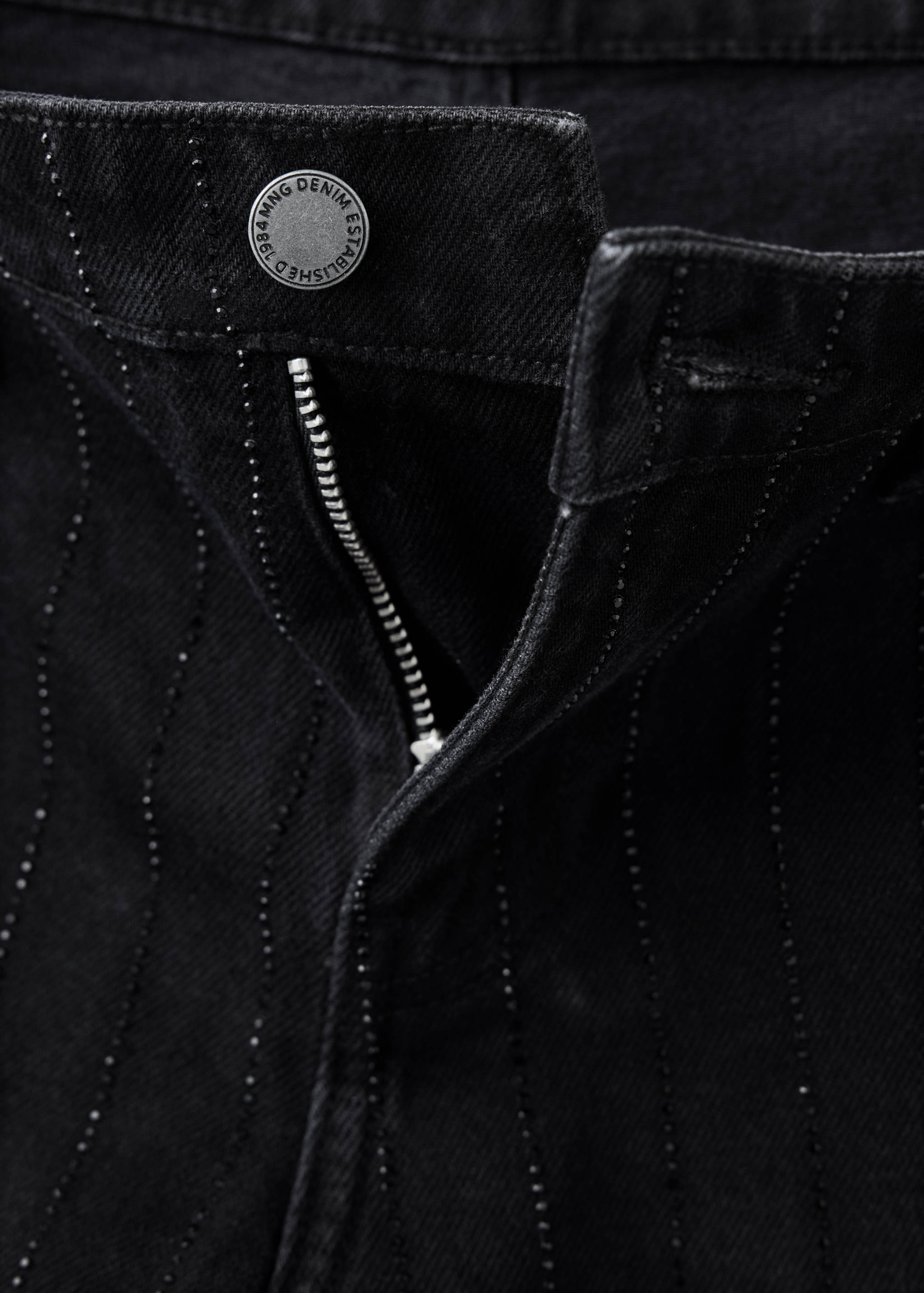 Primer plano de pantalones vaqueros negros con bragueta de botones y aplicaciones de pedrería verticales.