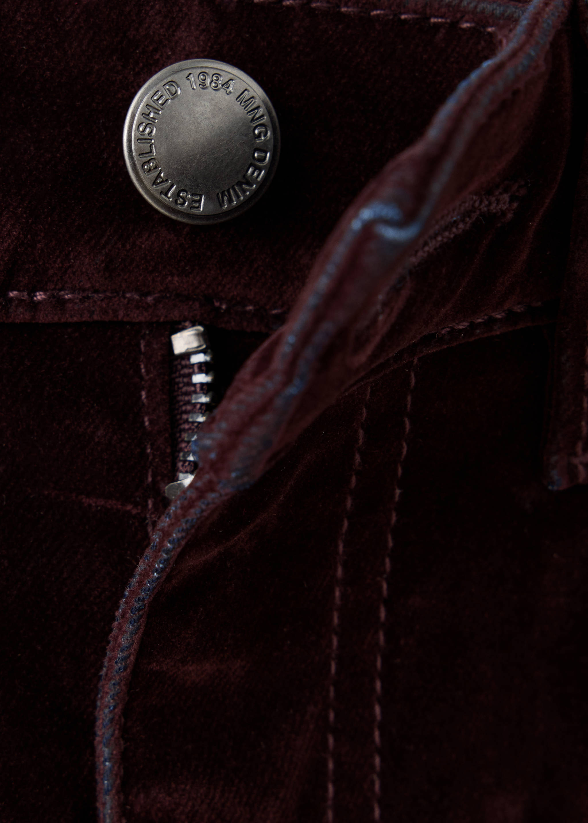 Detalle de pantalón de pana en color burdeos que muestra el botón metálico y la bragueta.