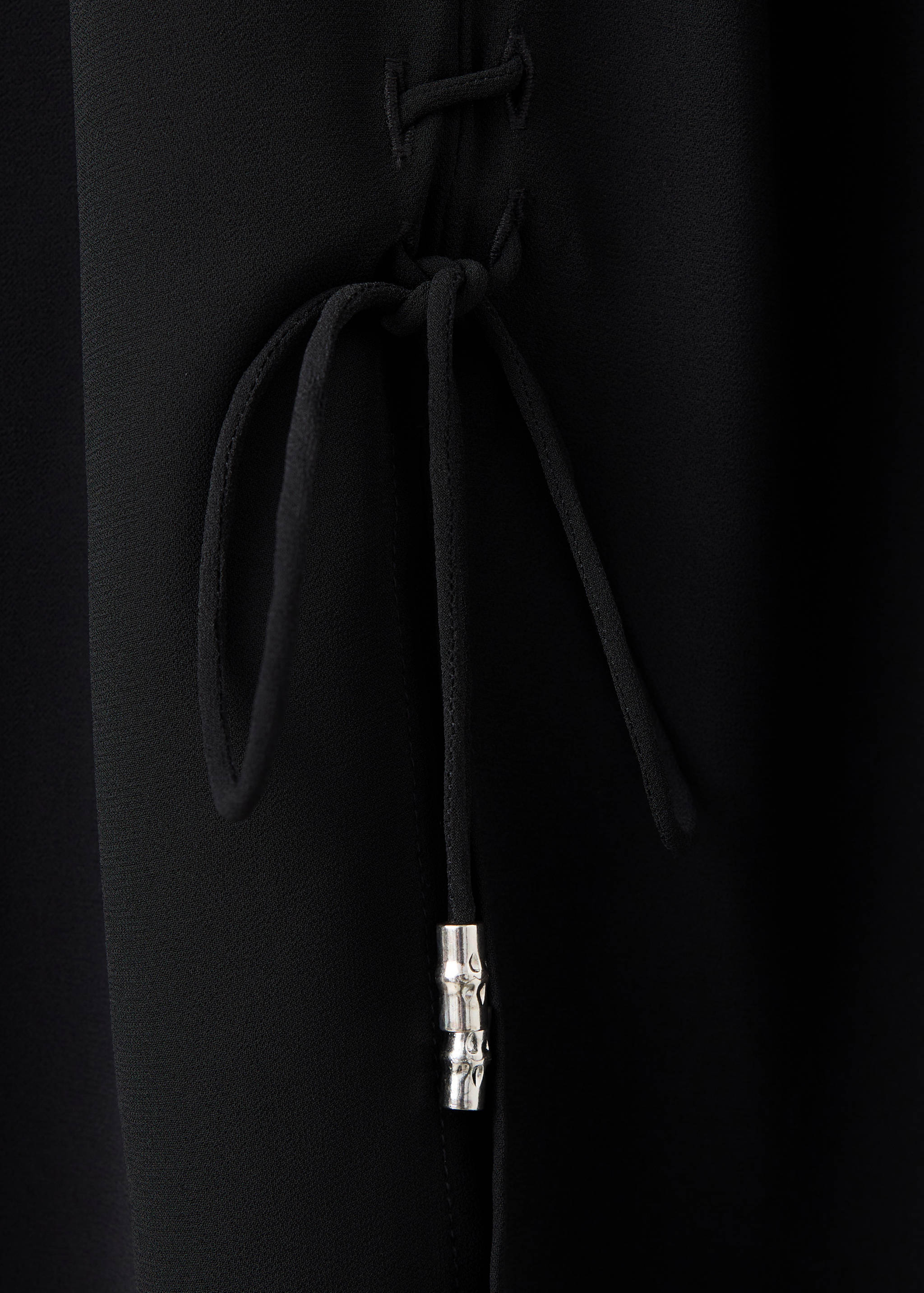 Primer plano de una blusa negra con lazada lateral y tiras con terminales metálicos.