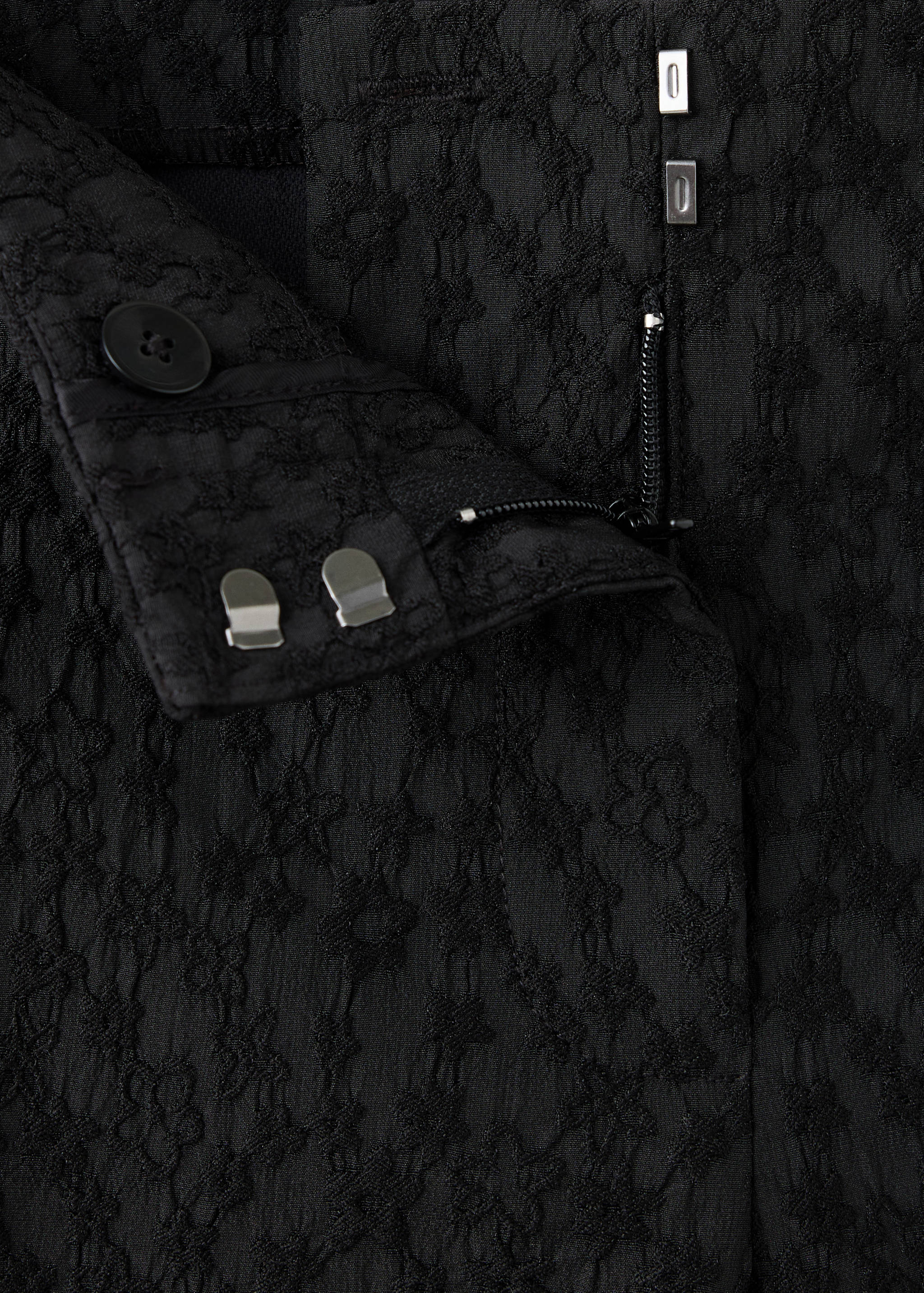 Close-up of black textured trousers showing the waistband and fastening details.