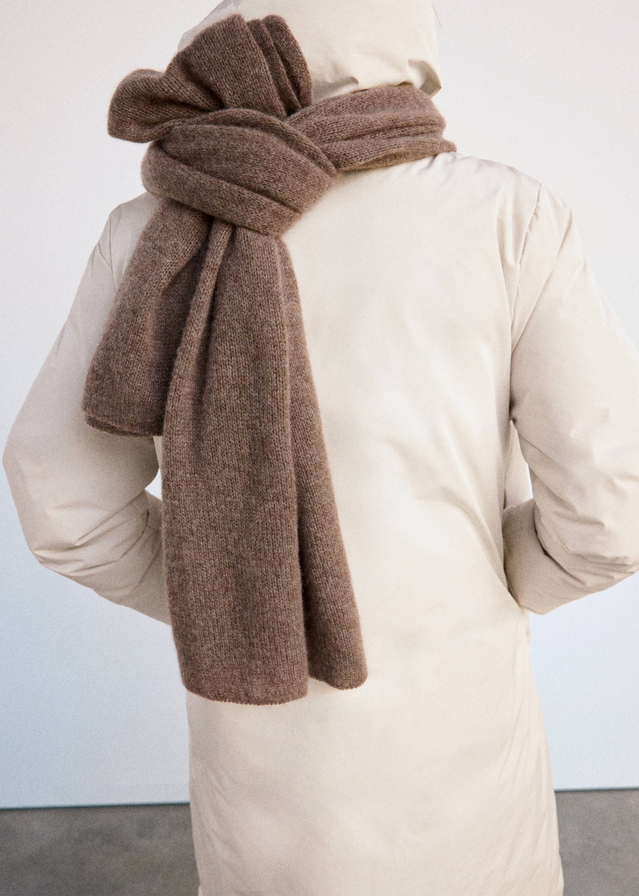 Back view of a beige anorak styled with a thick brown knitted scarf.