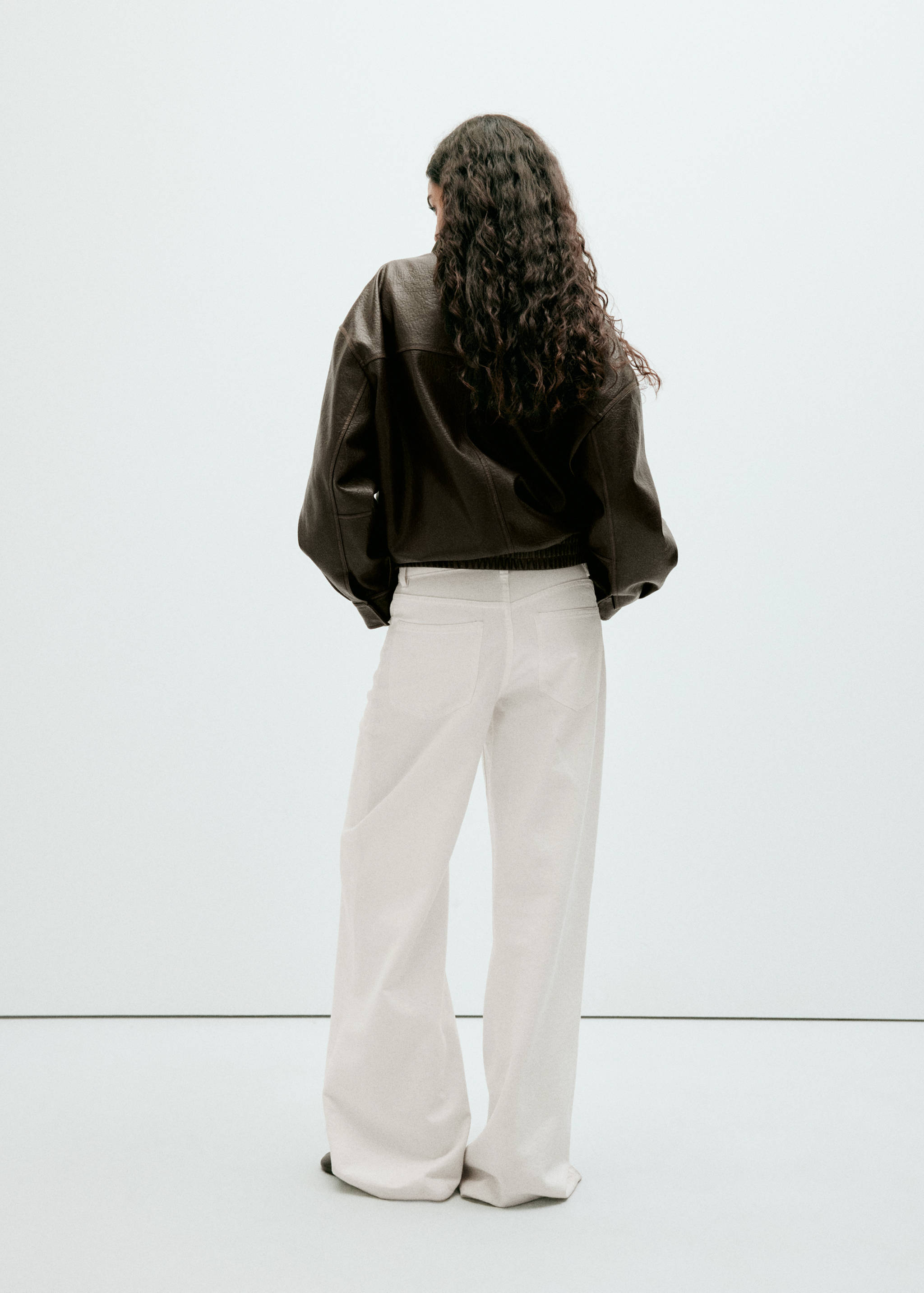 Back view of a dark brown leather jacket with a relaxed fit, worn with white trousers.