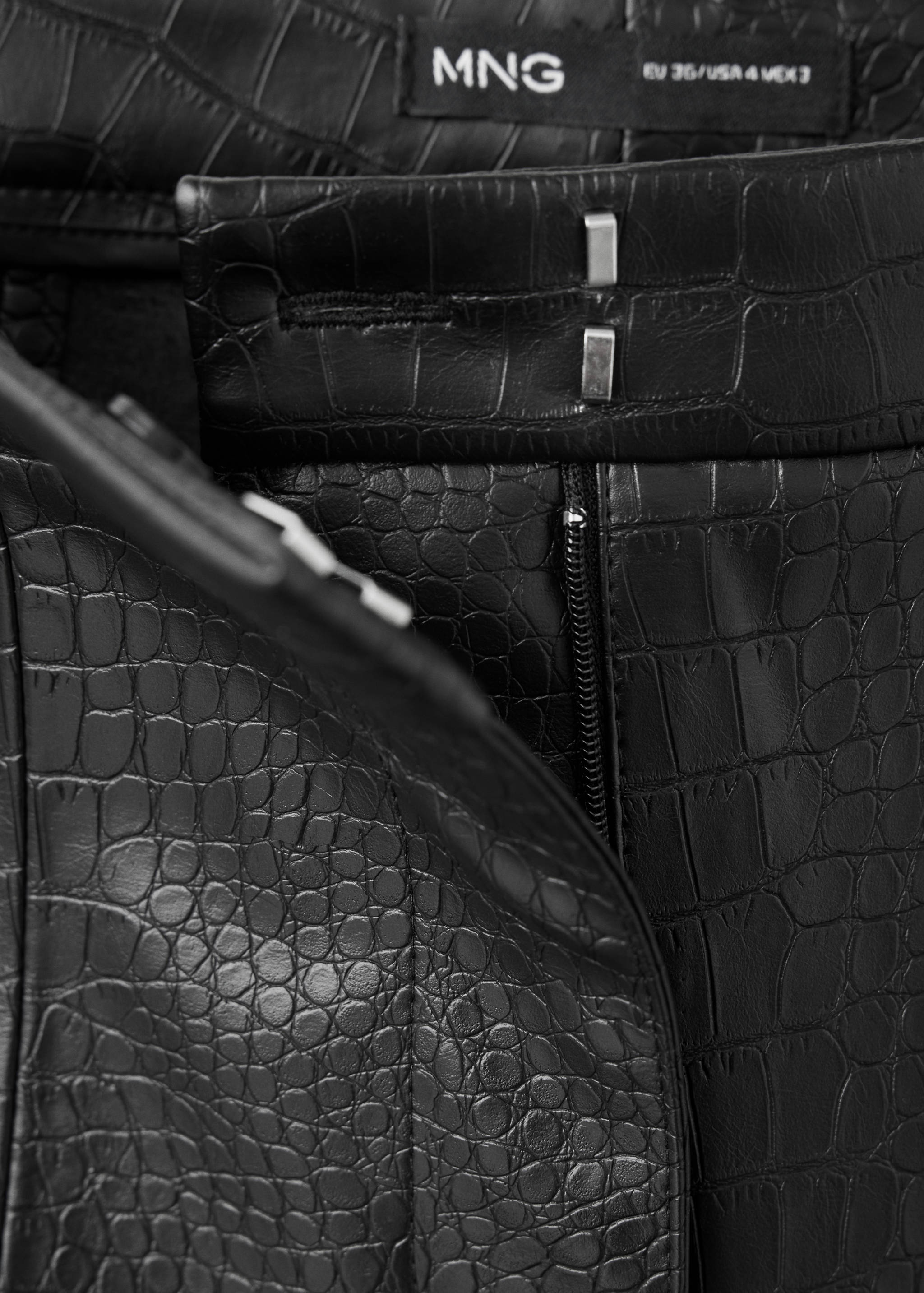 Close-up of black crocodile-textured trousers waistband and fastening detail.