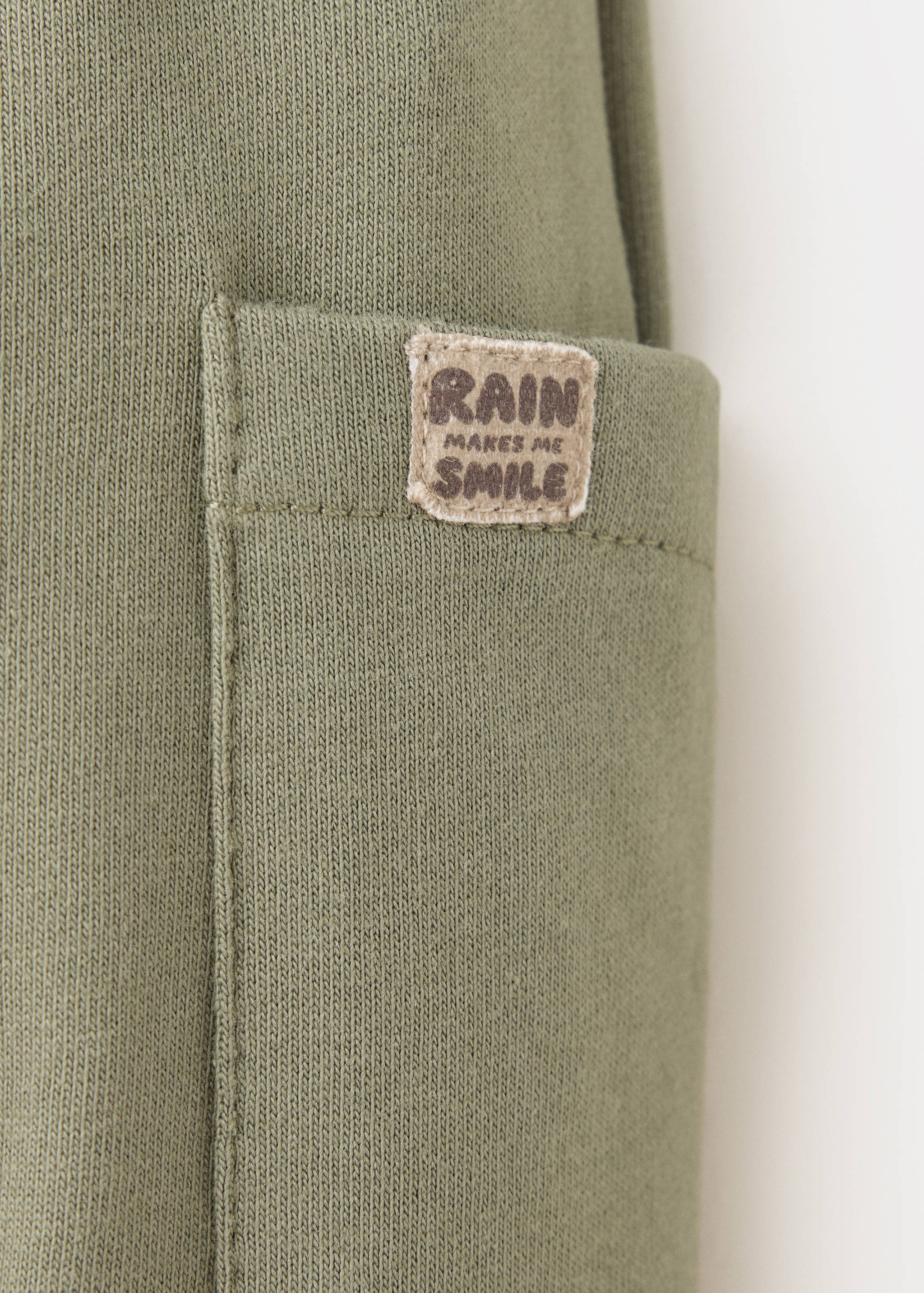 Close-up of khaki green joggers pocket with 'Rain Makes Me Smile' label.