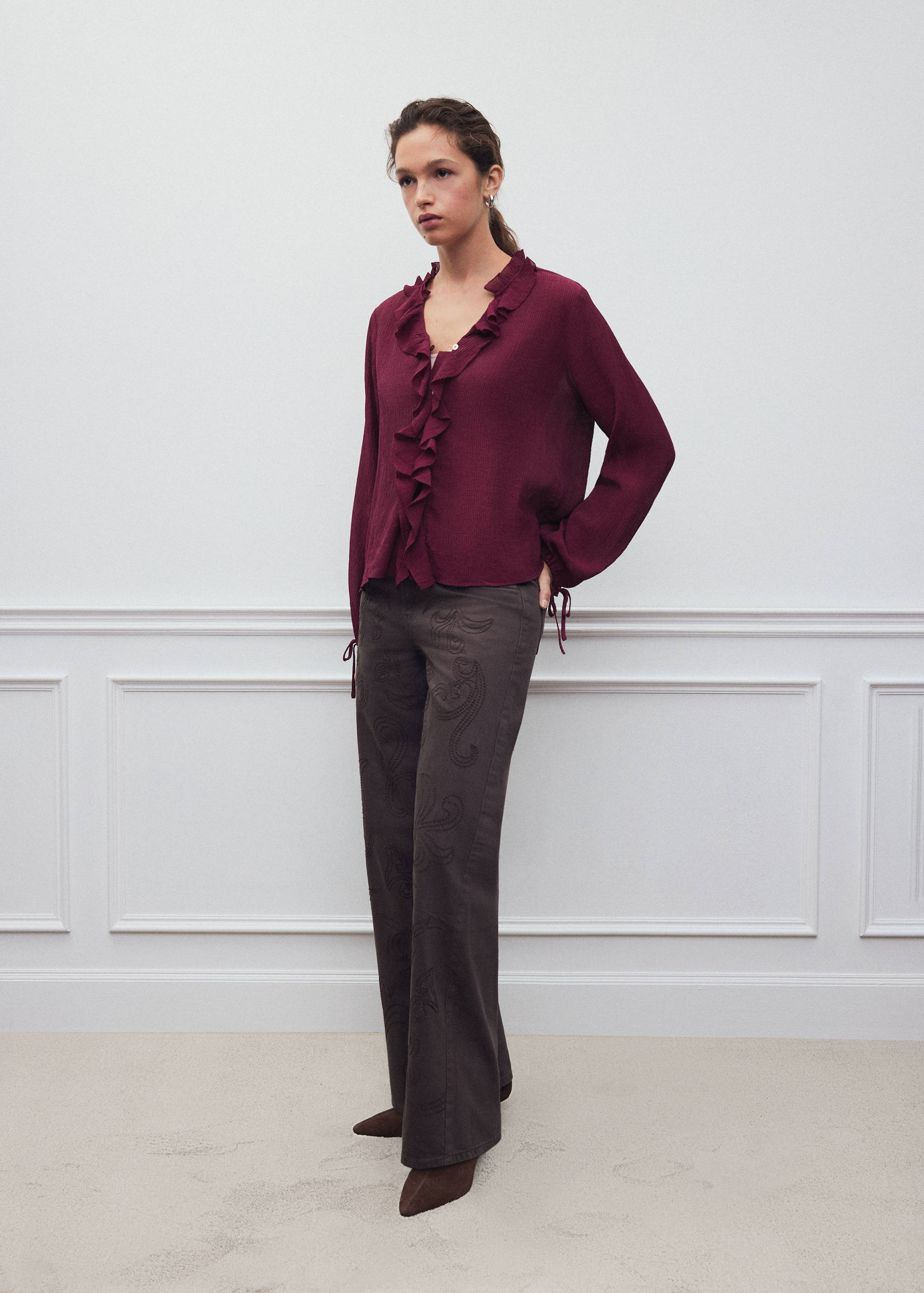 Burgundy long-sleeved shirt with ruffled trim and cuff ties, styled with dark trousers.