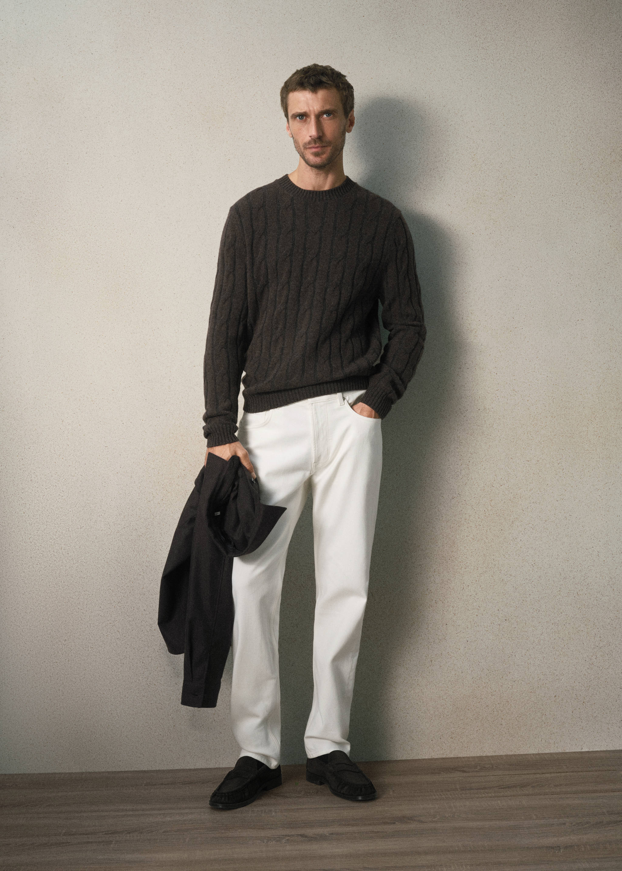 Dark brown cable-knit jersey with crew neck, worn with white trousers.