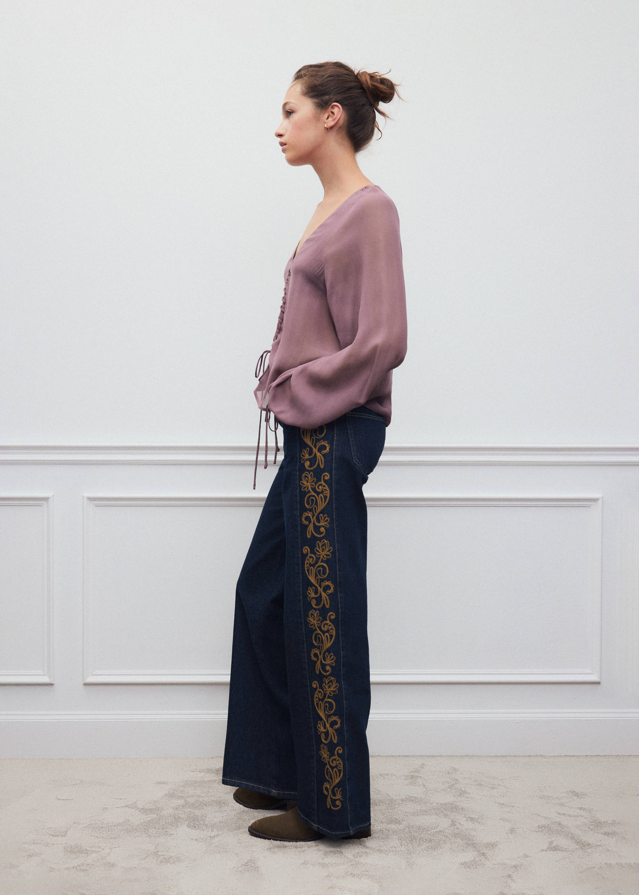Pantalones oscuros con lavado y bordados con motivo floral lateral, combinados con una blusa malva.