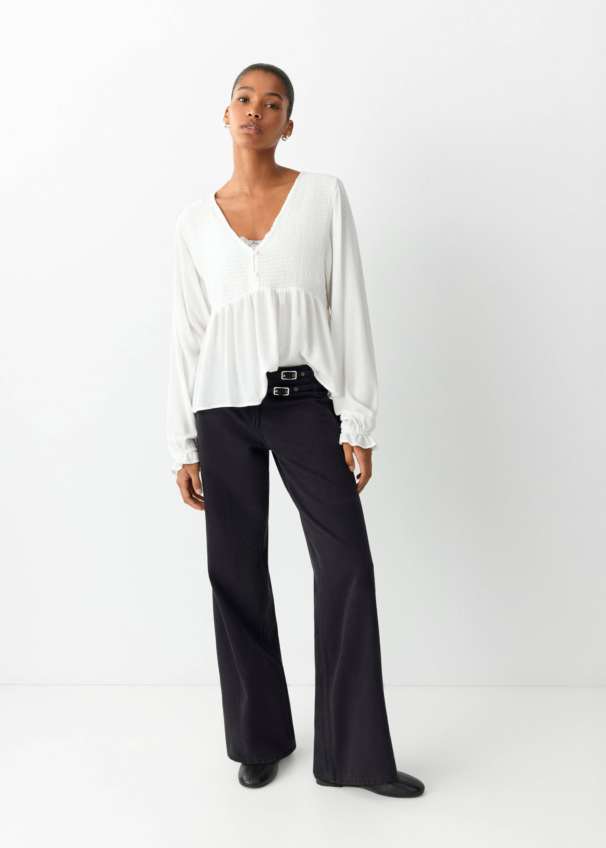 White long-sleeved V-neck shirt with a smocked bodice, worn with black trousers.