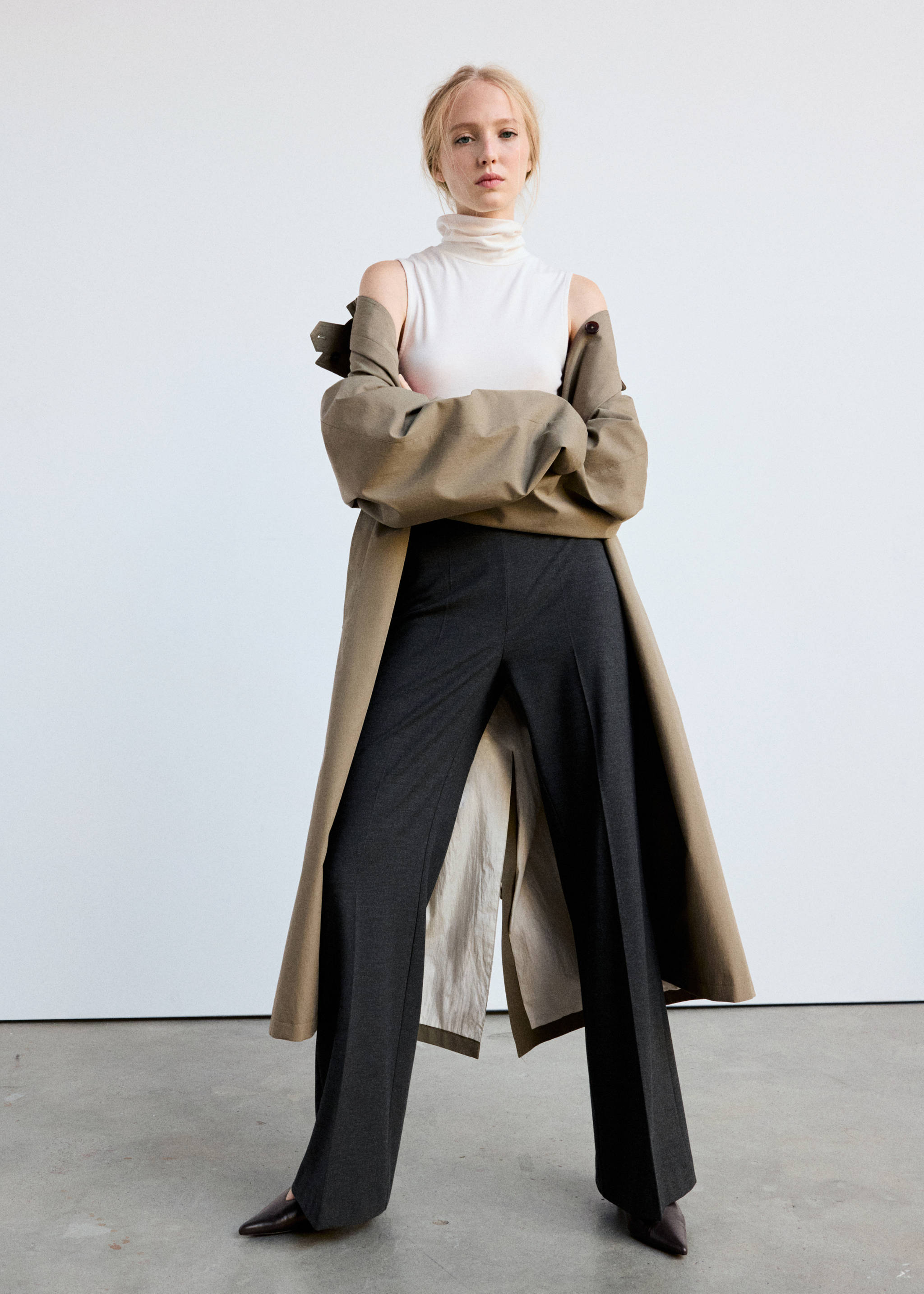 Dark grey trousers with a pressed front crease, styled with a white top and olive coat.