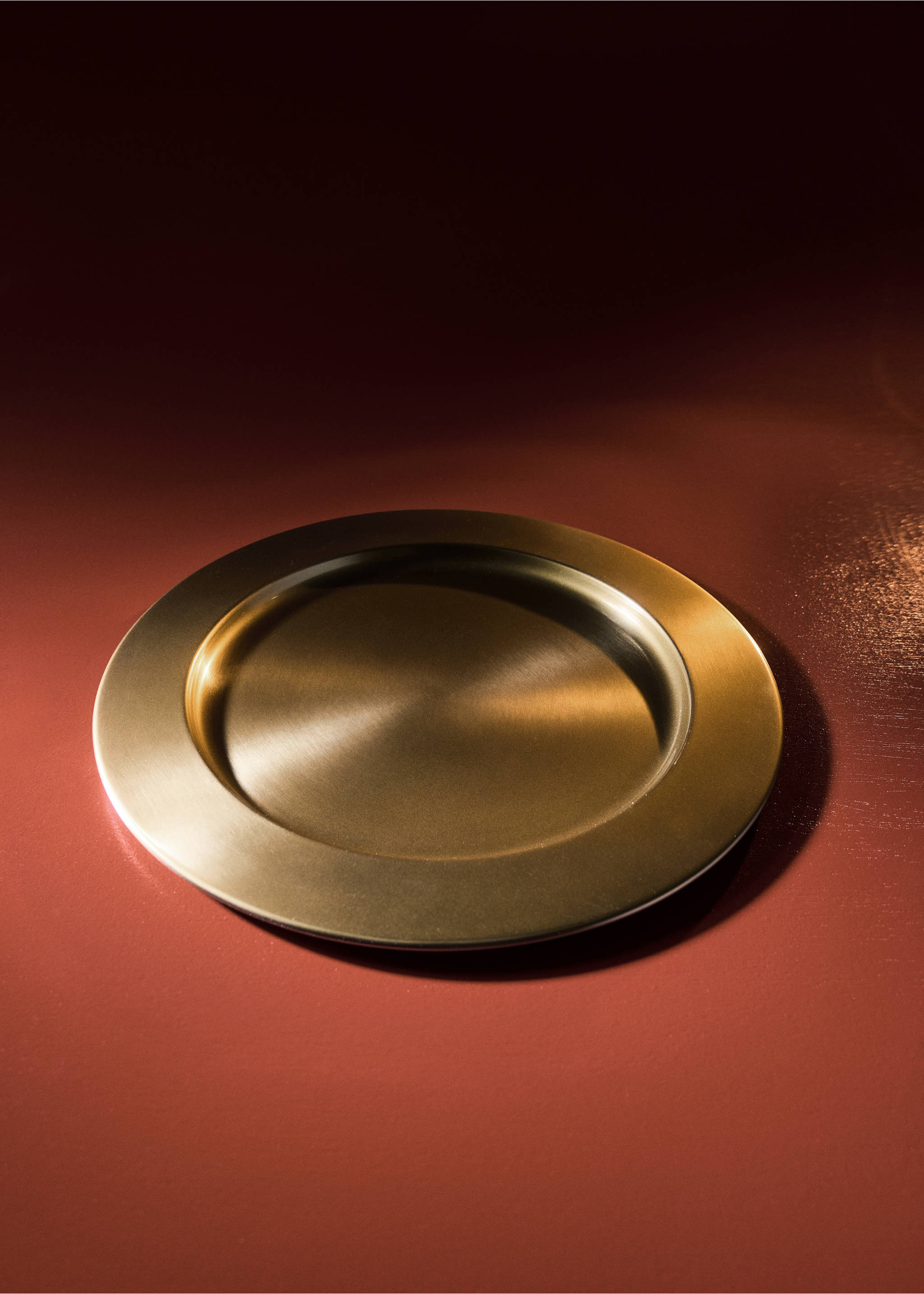A round gold-toned brushed metal plate on a dark red surface., Gold. Ref: 37057893-00.