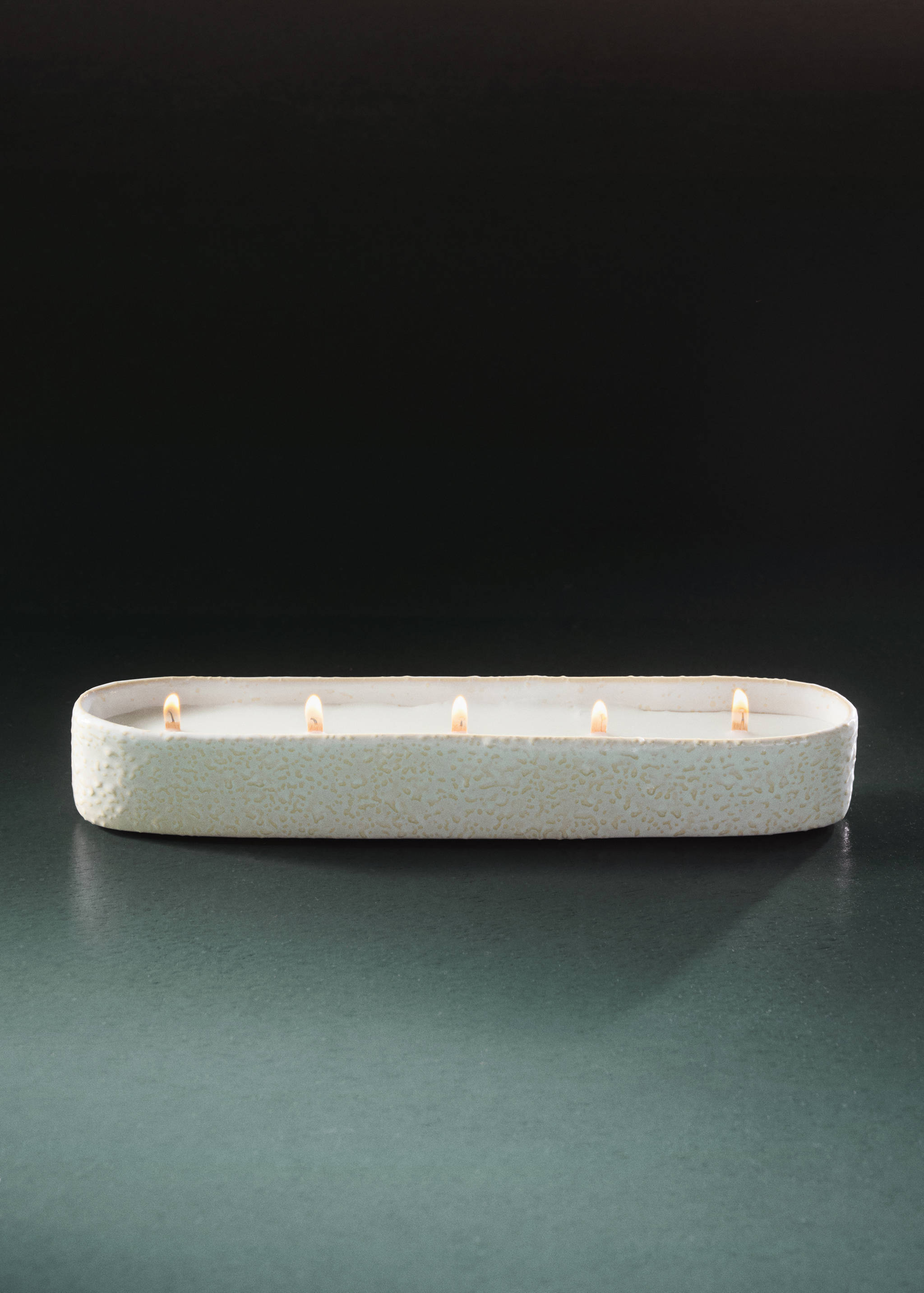 An elongated oval candle in a textured off-white container with five lit wicks.