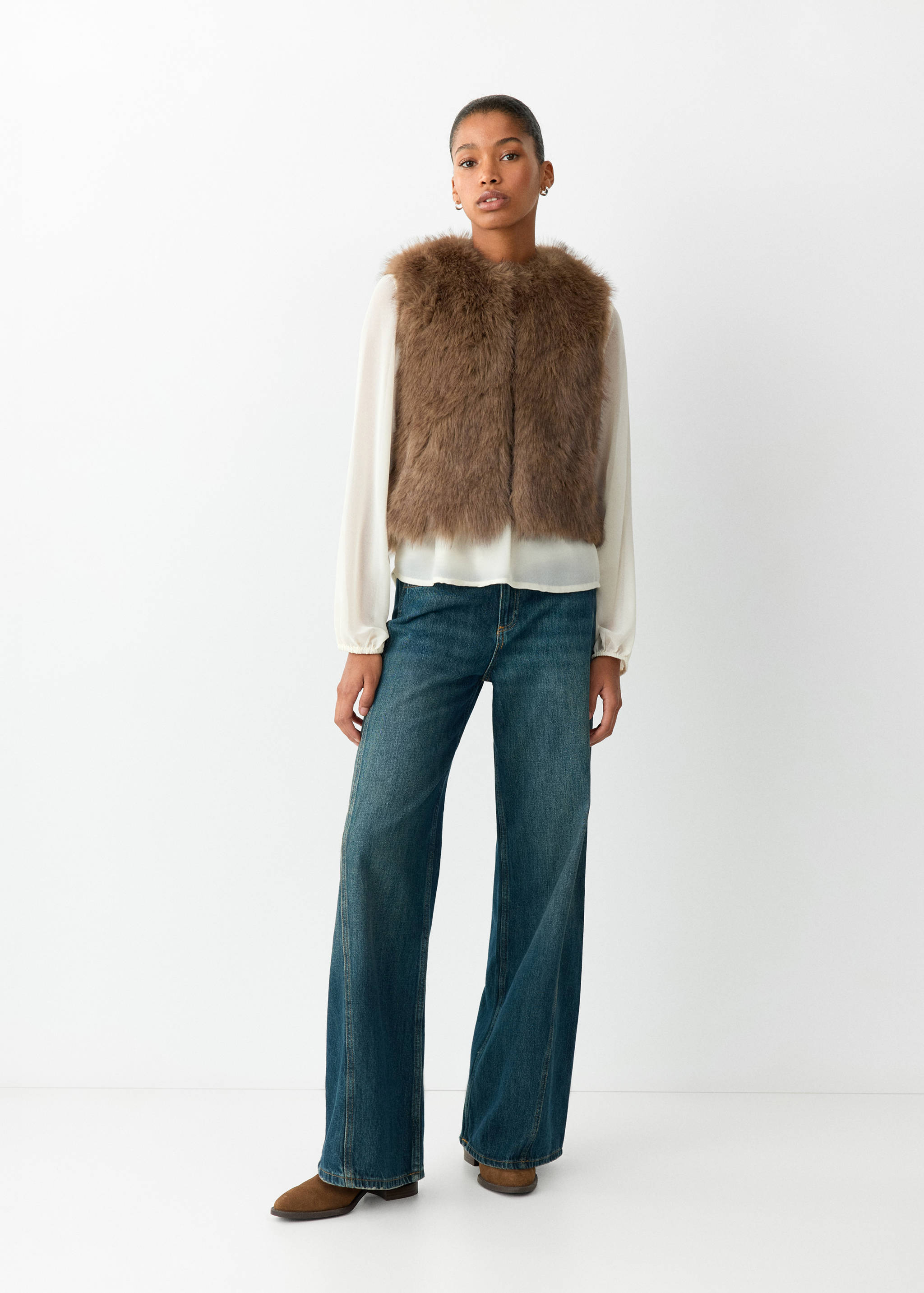 Brown textured faux-fur vest worn with a white blouse and blue denim trousers.