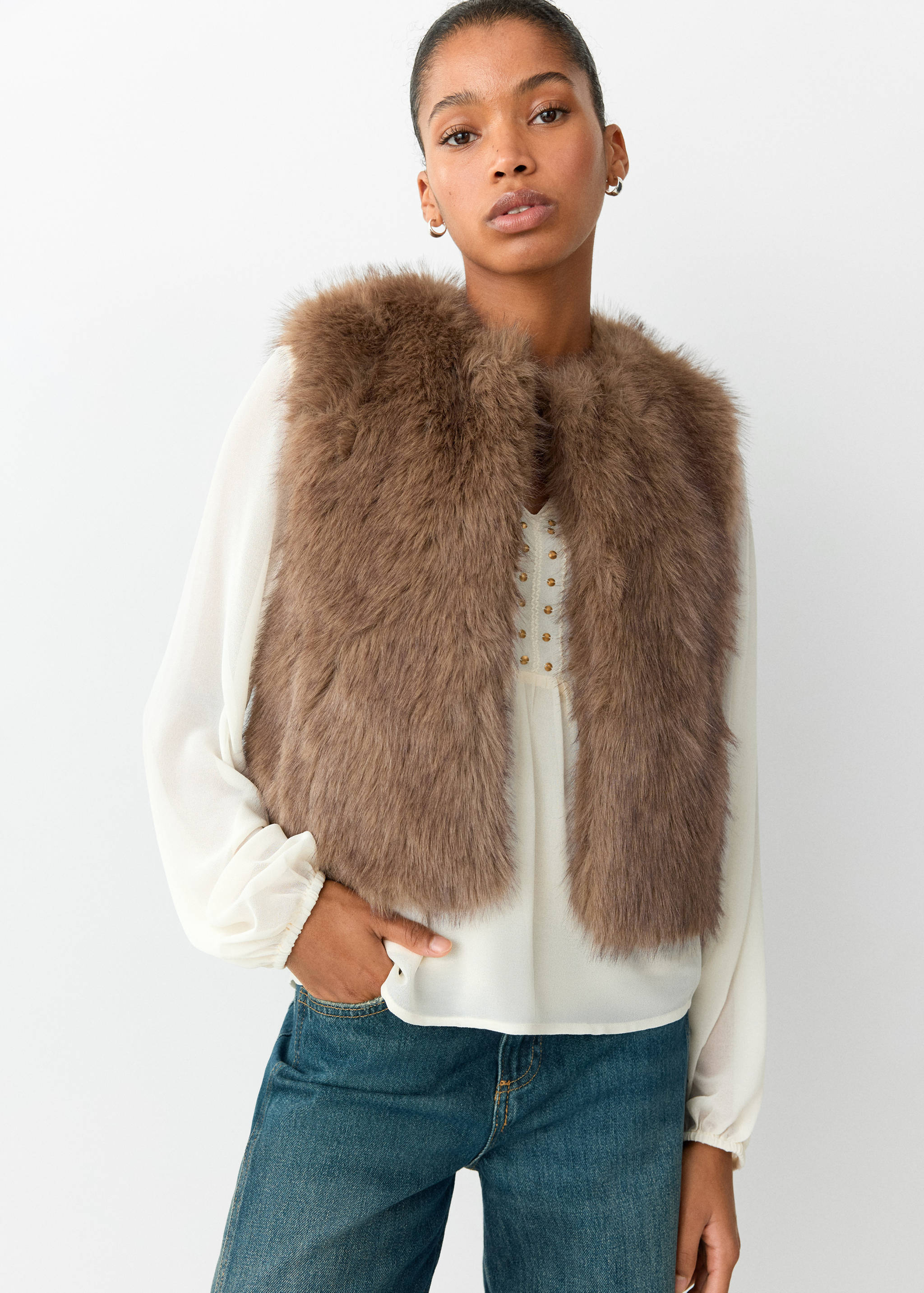 Brown faux-fur vest worn over a cream blouse and blue denim trousers.