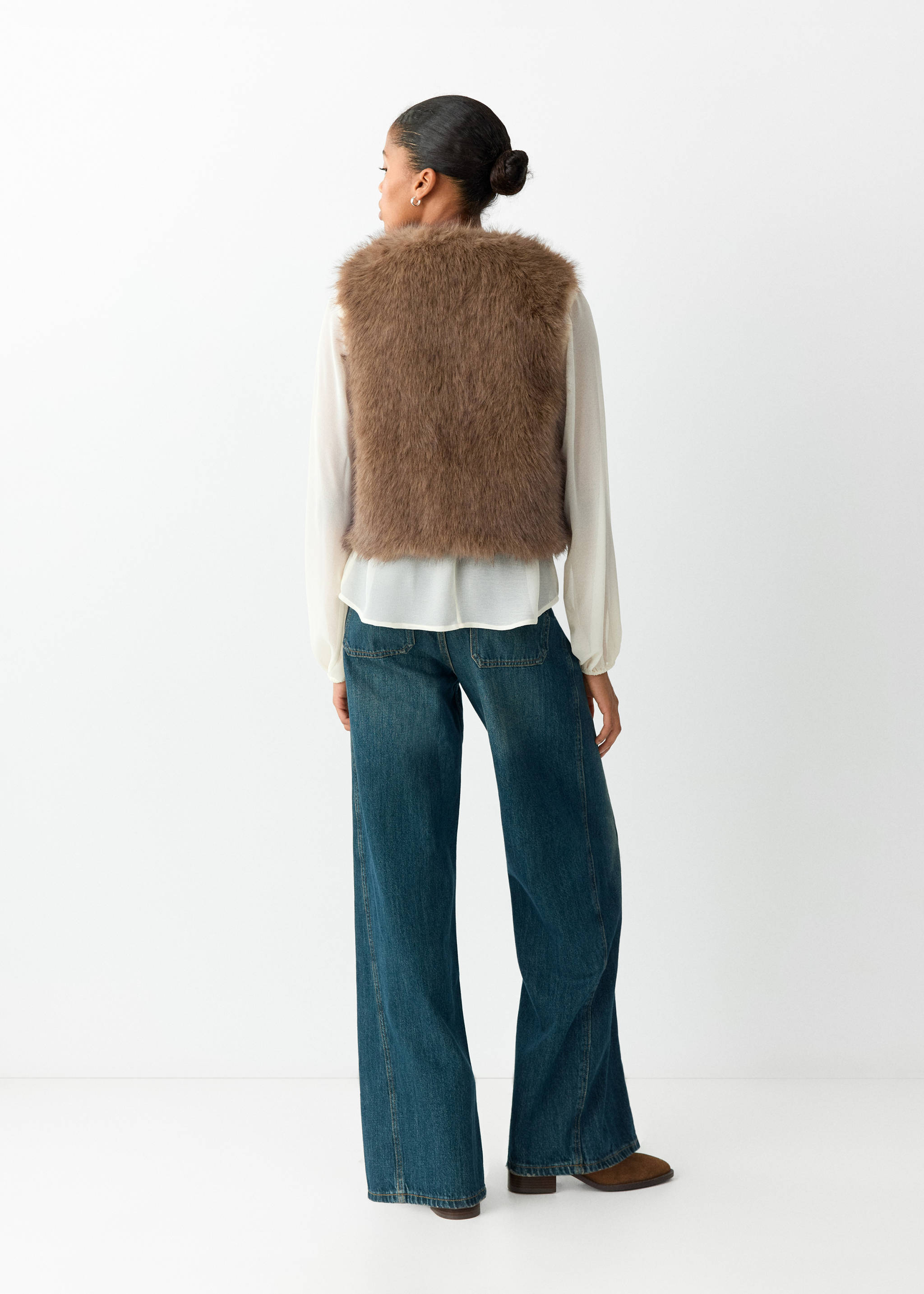 Brown faux-fur vest shown from the back, layered over a white blouse and blue trousers.