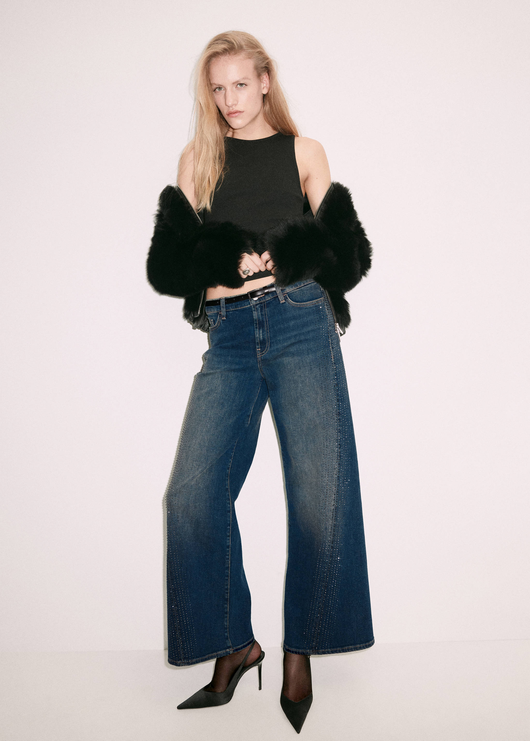 Pantalones vaqueros anchos azules con apliques de pedrería en los laterales, combinados con top negro y chaqueta efecto pelo.