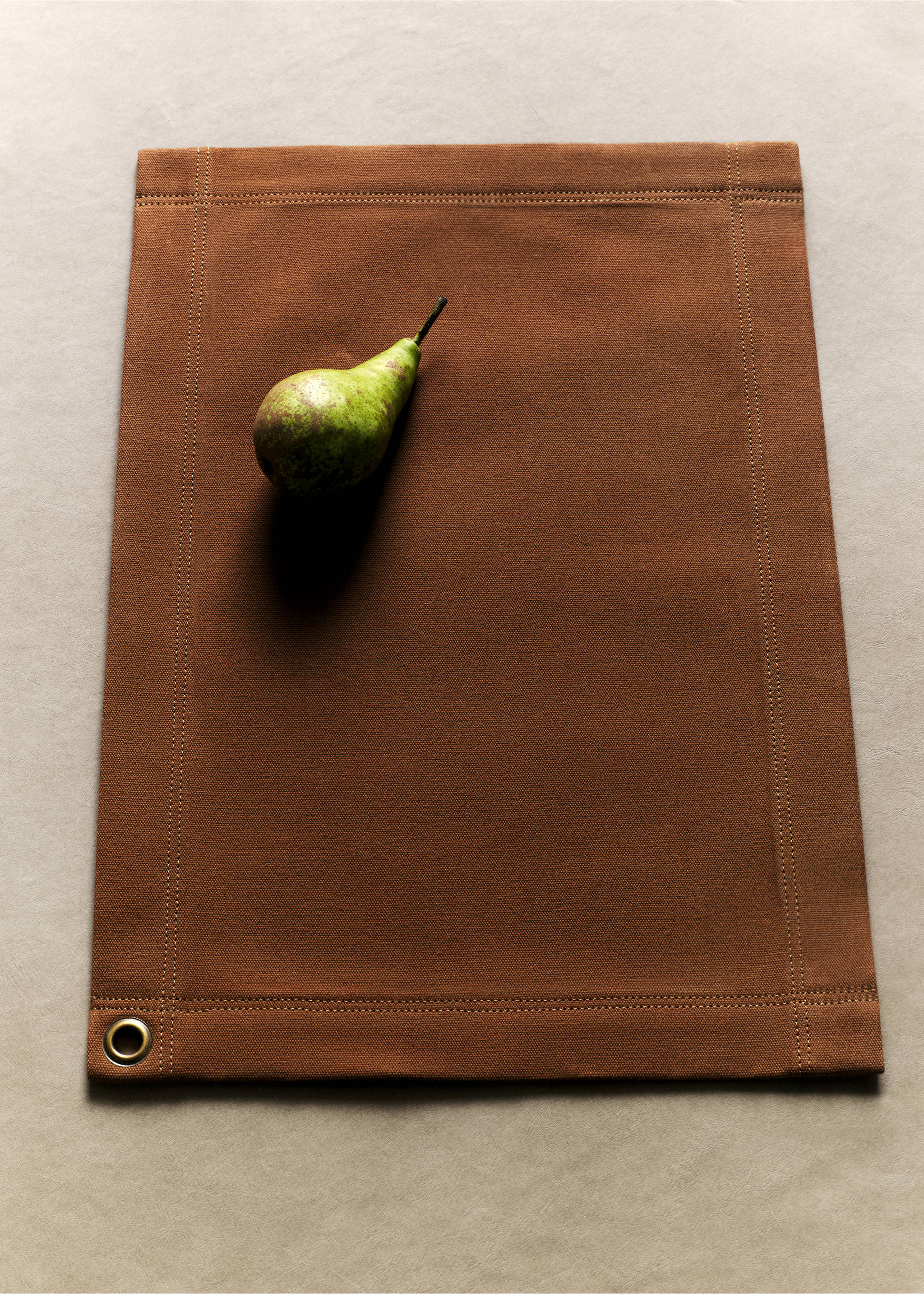 Brown placemat with topstitching and a corner eyelet, styled with a pear., Brown. Ref: 37062542-00.